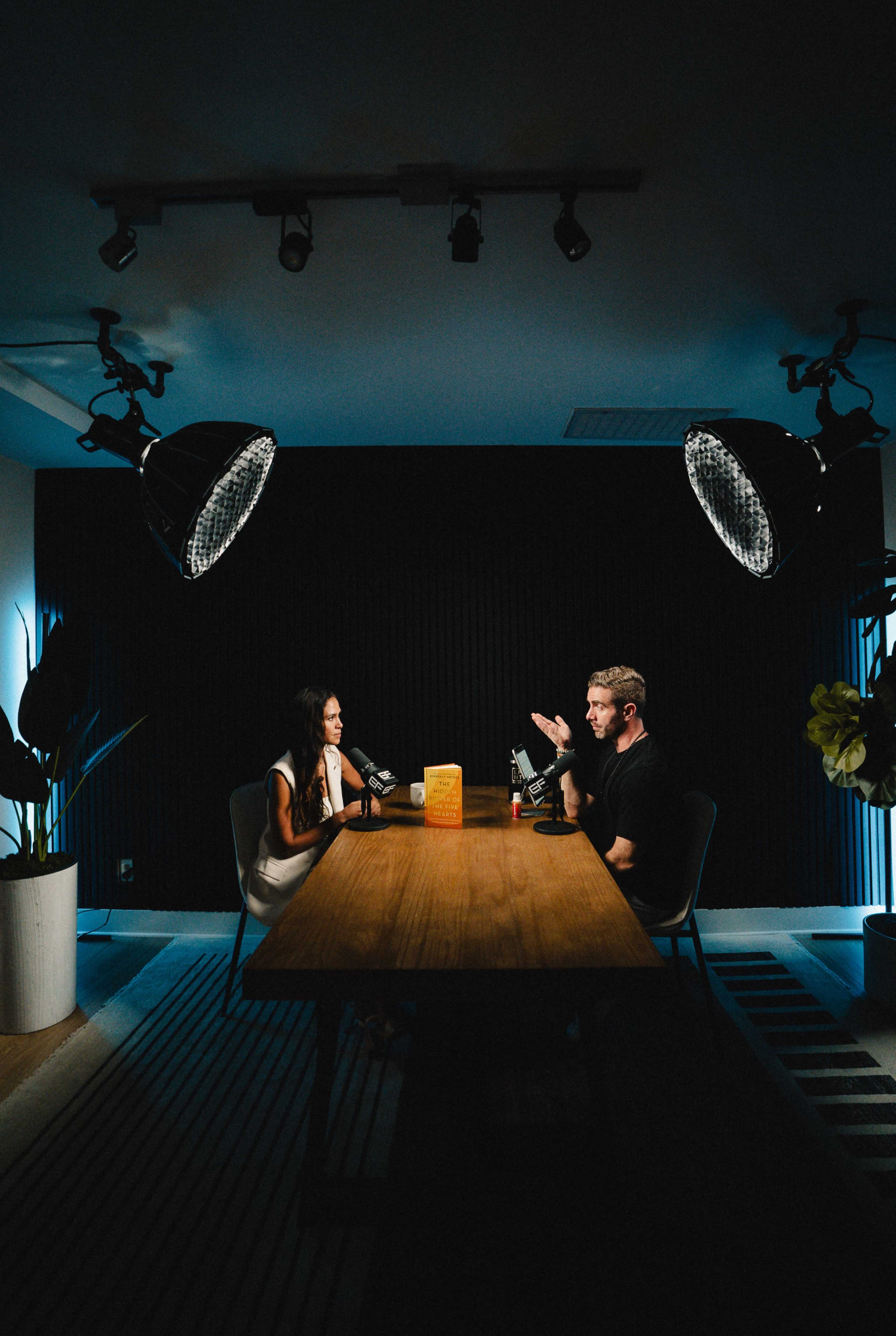 Two people are seated at a wooden table in a darkly lit room, with large studio lights illuminating them as they engage in a conversation, surrounded by plants.