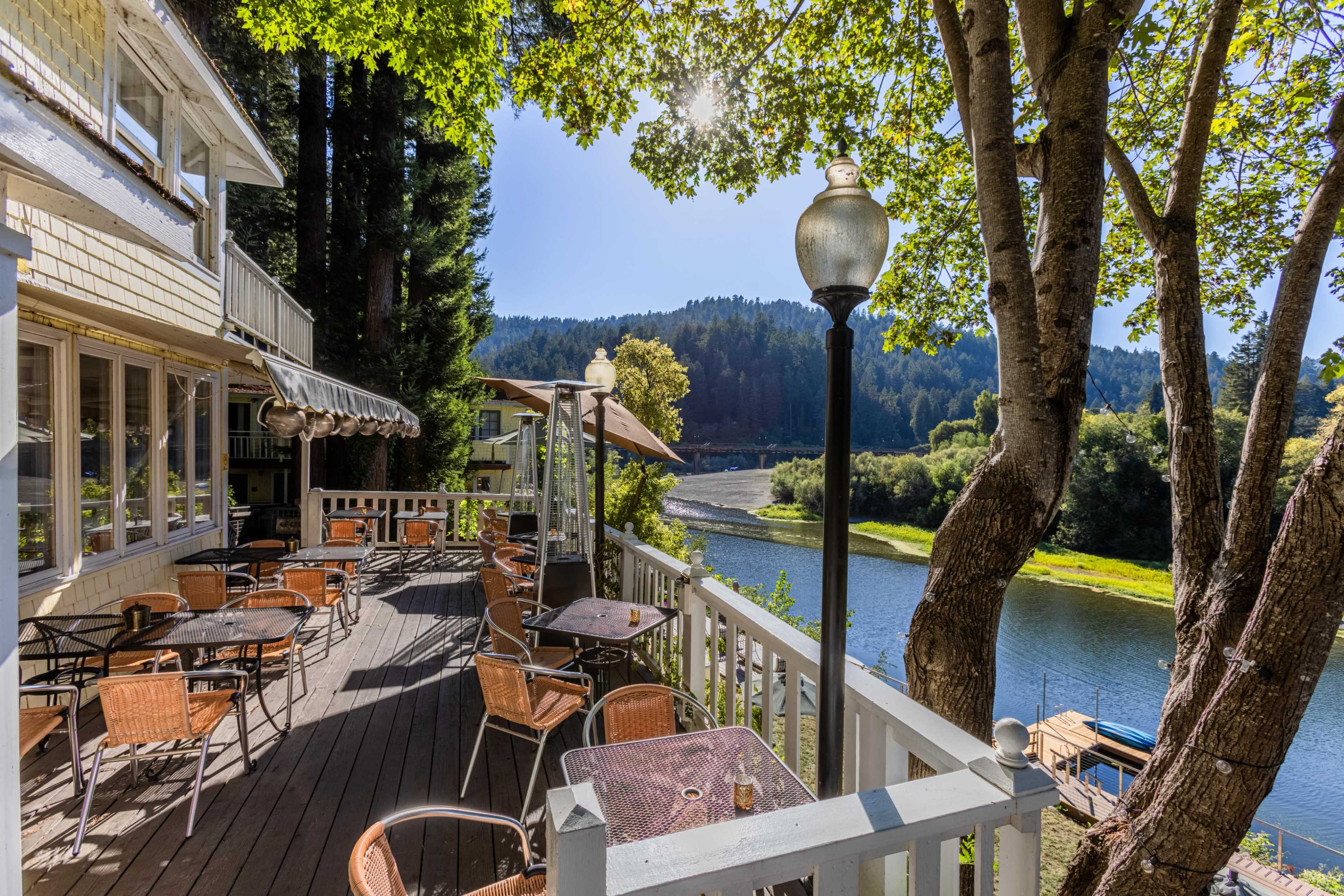 A riverside deck with empty tables and chairs overlooks a calm river surrounded by trees and hills.
