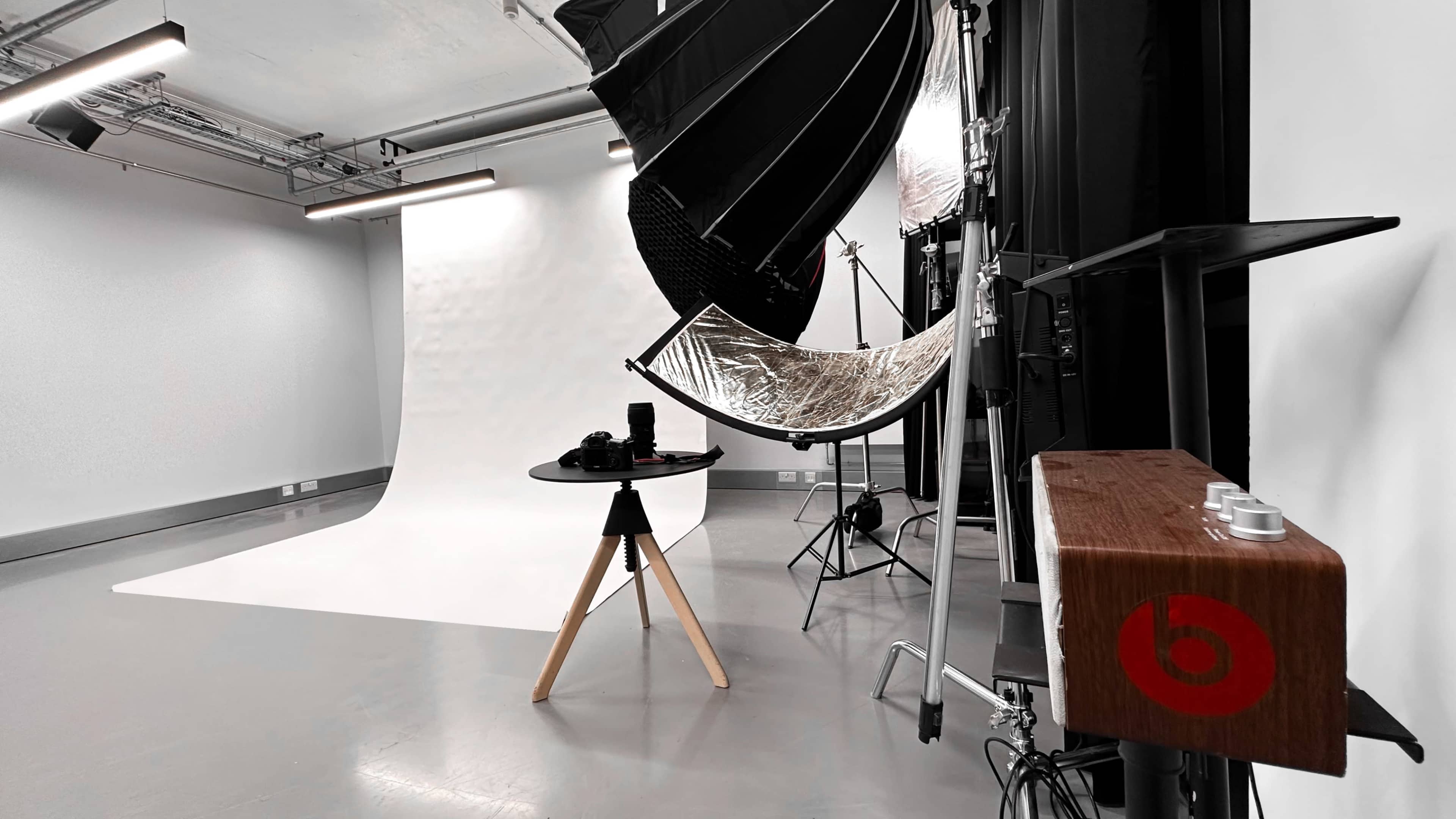 A photography studio setup with a white backdrop, lighting equipment, and a table holding camera gear.