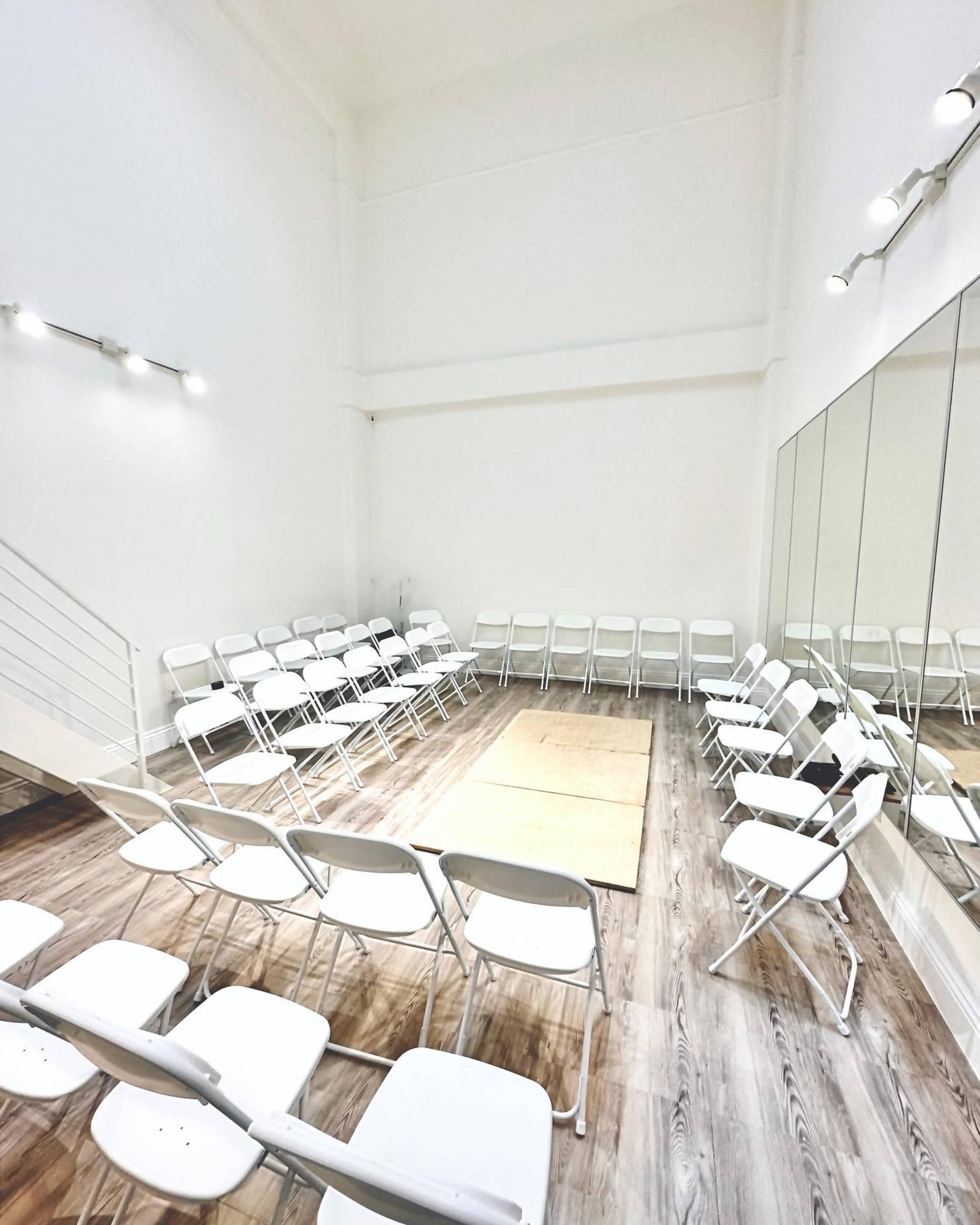 A group of white folding chairs is arranged in a circle around a small area on a wooden floor in a well-lit room with white walls and a large mirror.