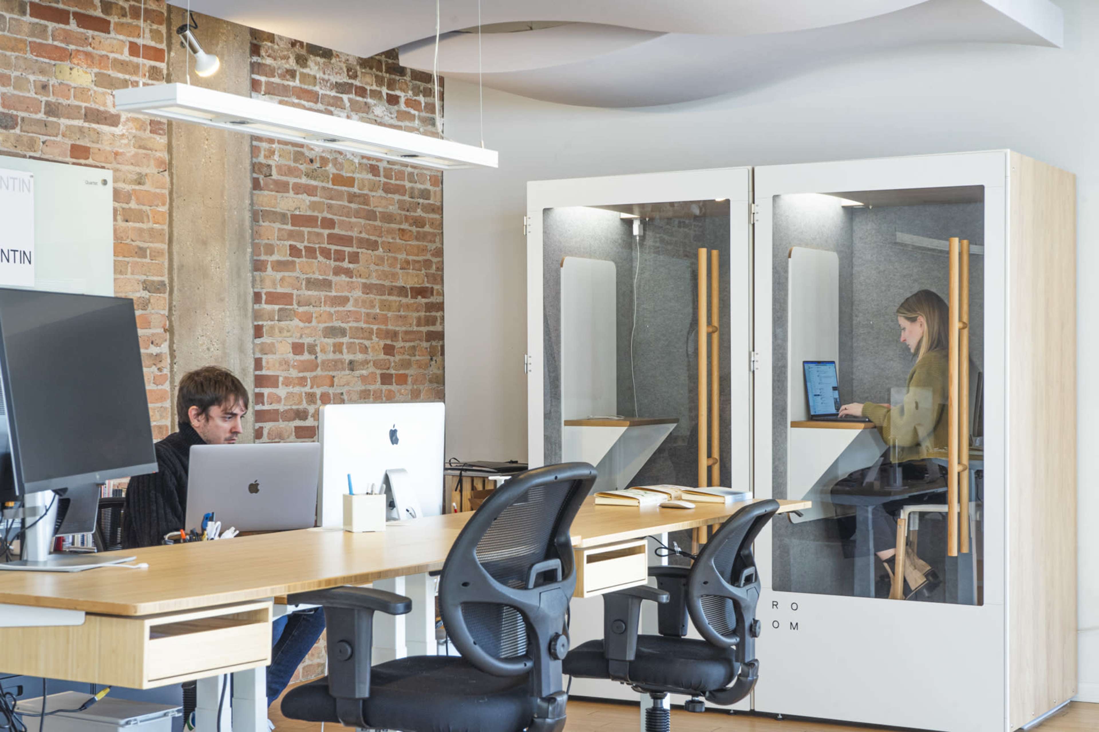 A modern office space features a wooden desk with two computers, one occupied by a man, while a woman works in a glass-walled pod nearby.