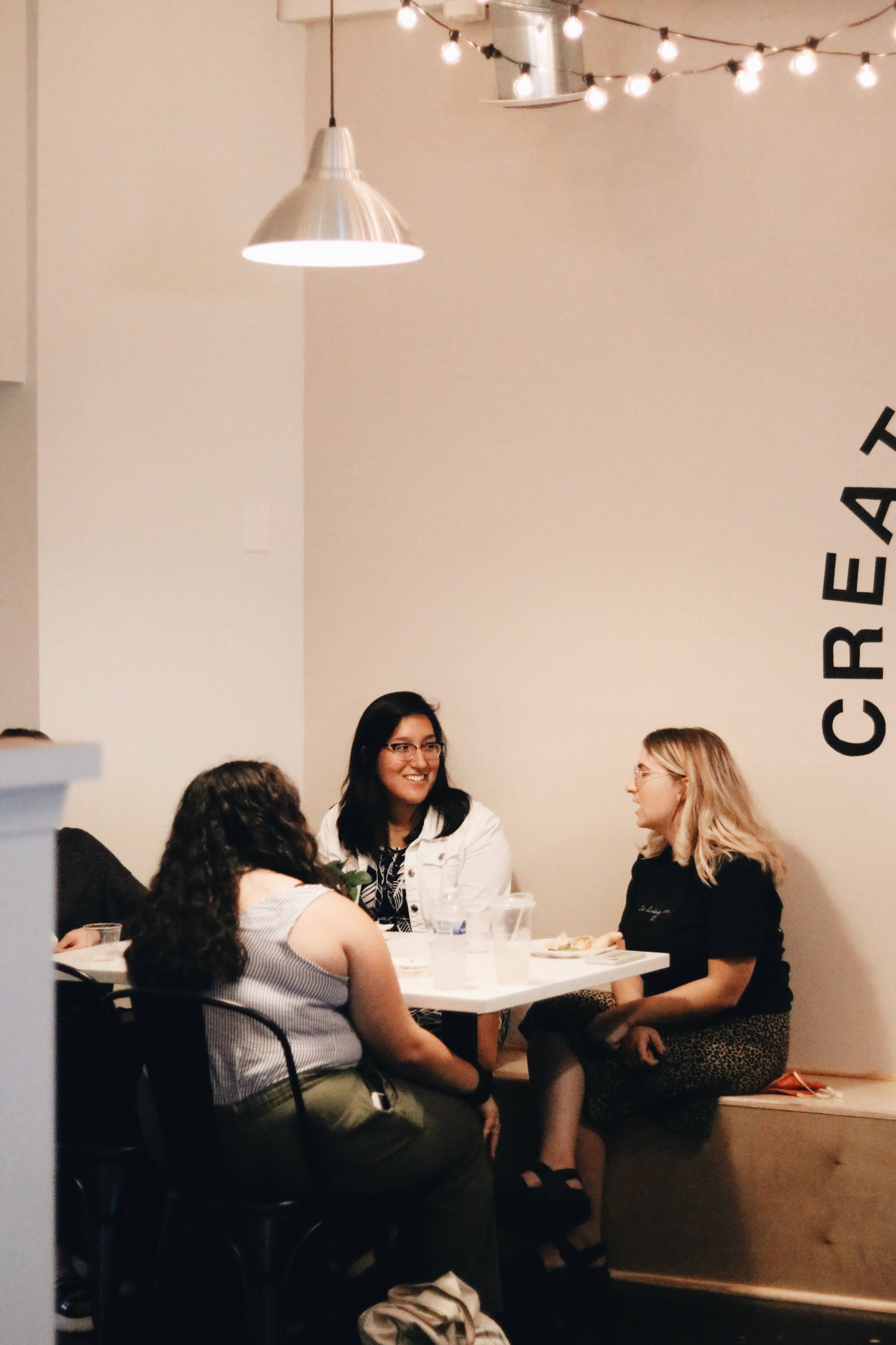 A group of people is seated at a table in a bright room, engaged in conversation near a wall with the word "CREATIVE" displayed.