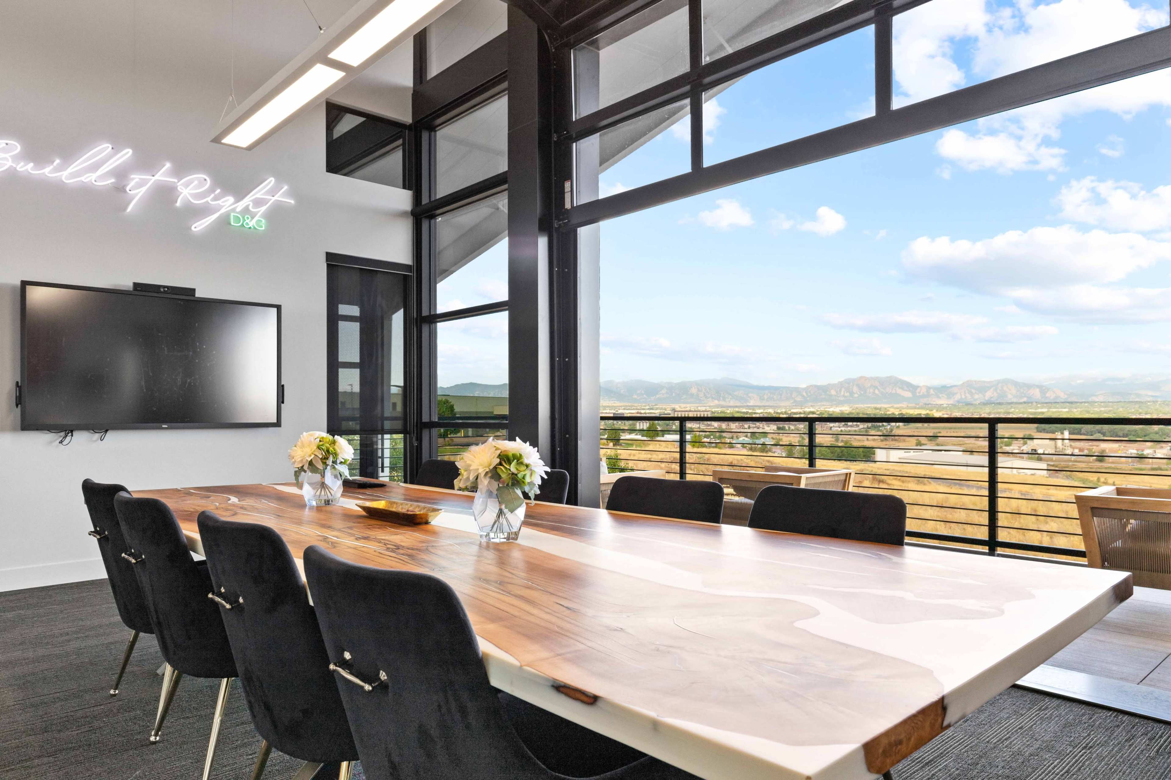 A modern conference room features a long wooden table, black chairs, and large windows with a view of mountains and the sky.