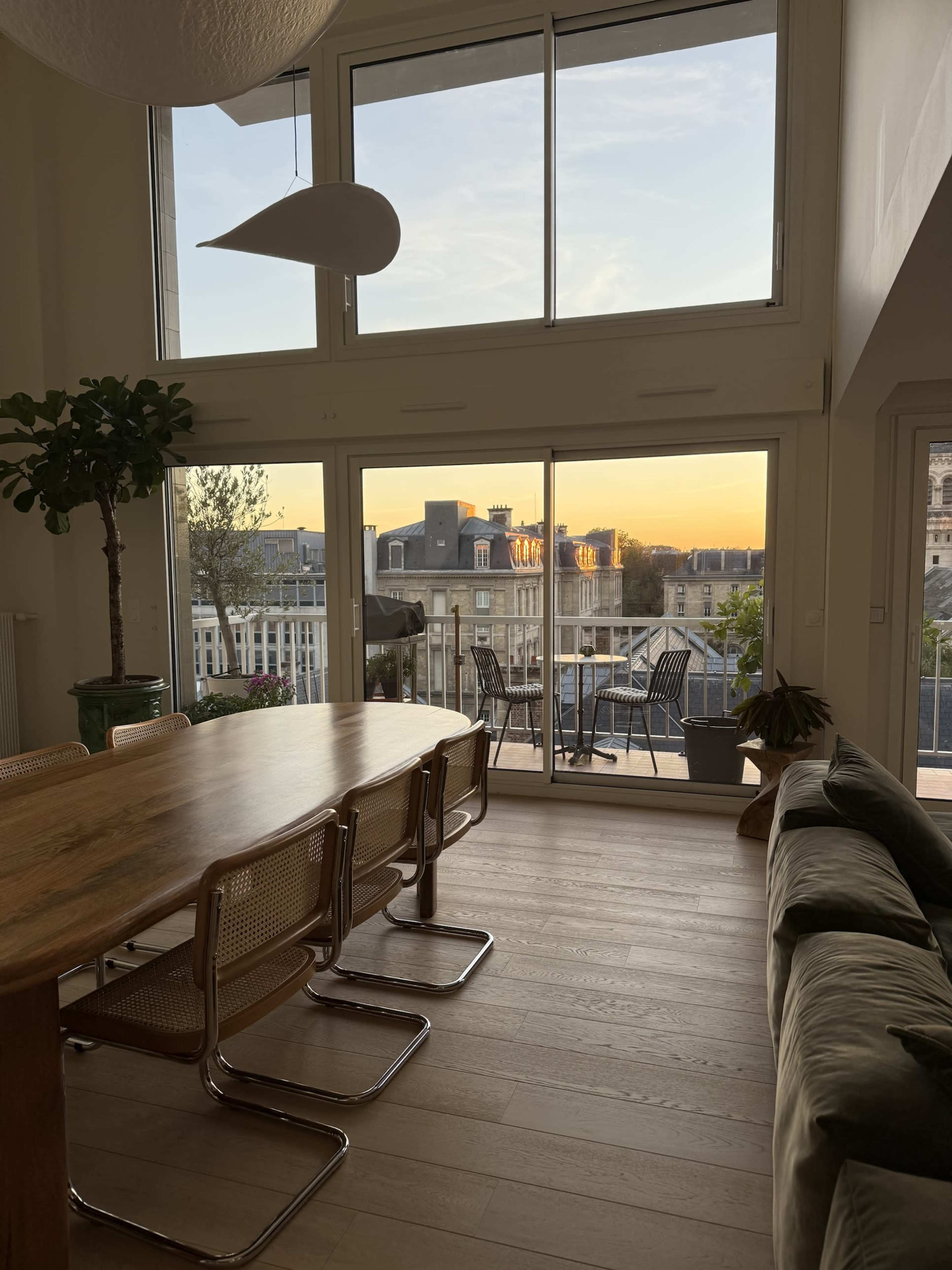 The image shows a bright interior with a large wooden dining table and chairs, facing a large glass door that opens to a balcony overlooking a sunset.