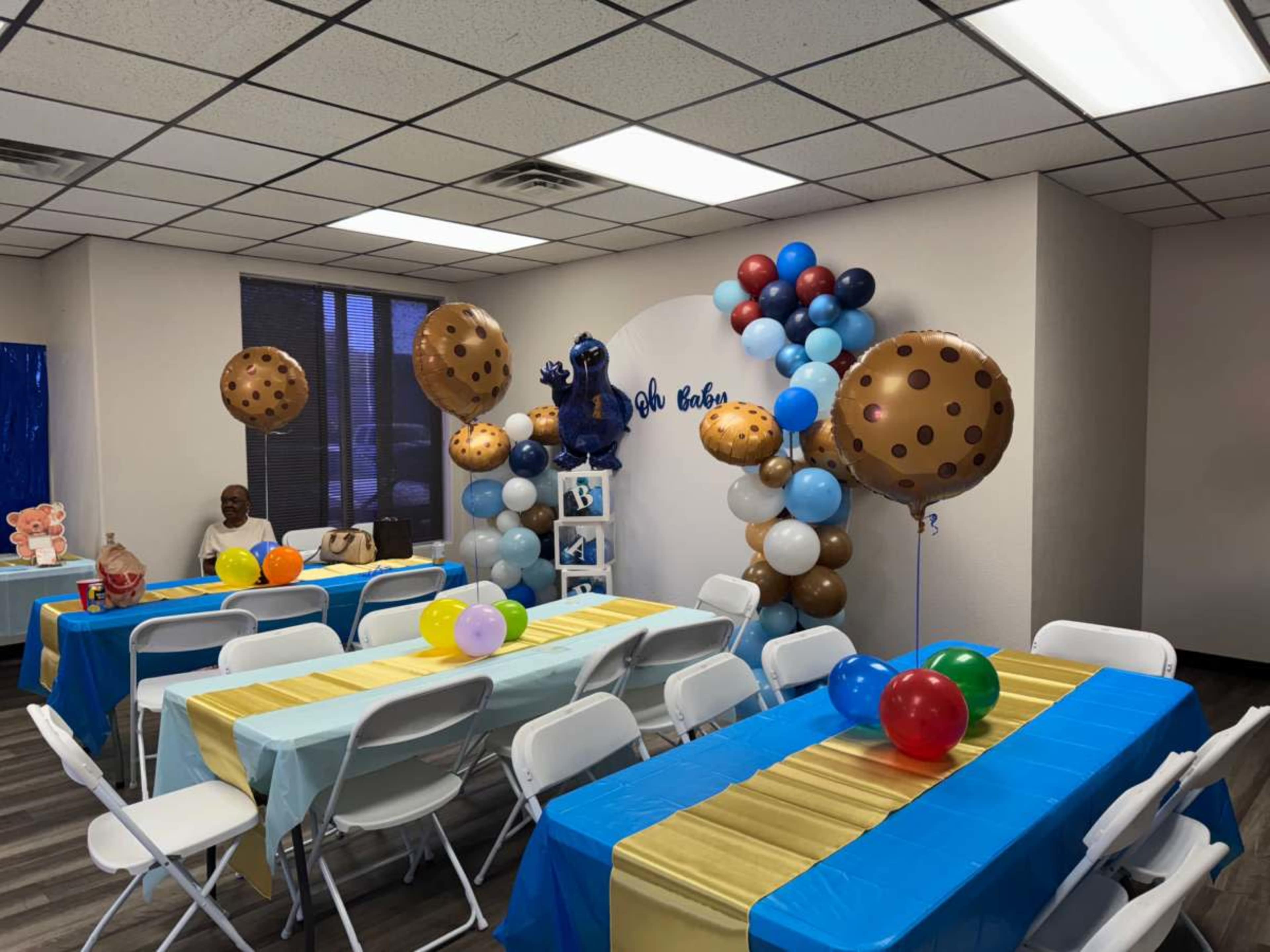 The image shows a decorated party room with blue and yellow tables, cookie-themed balloons, and a backdrop featuring a bear figure.