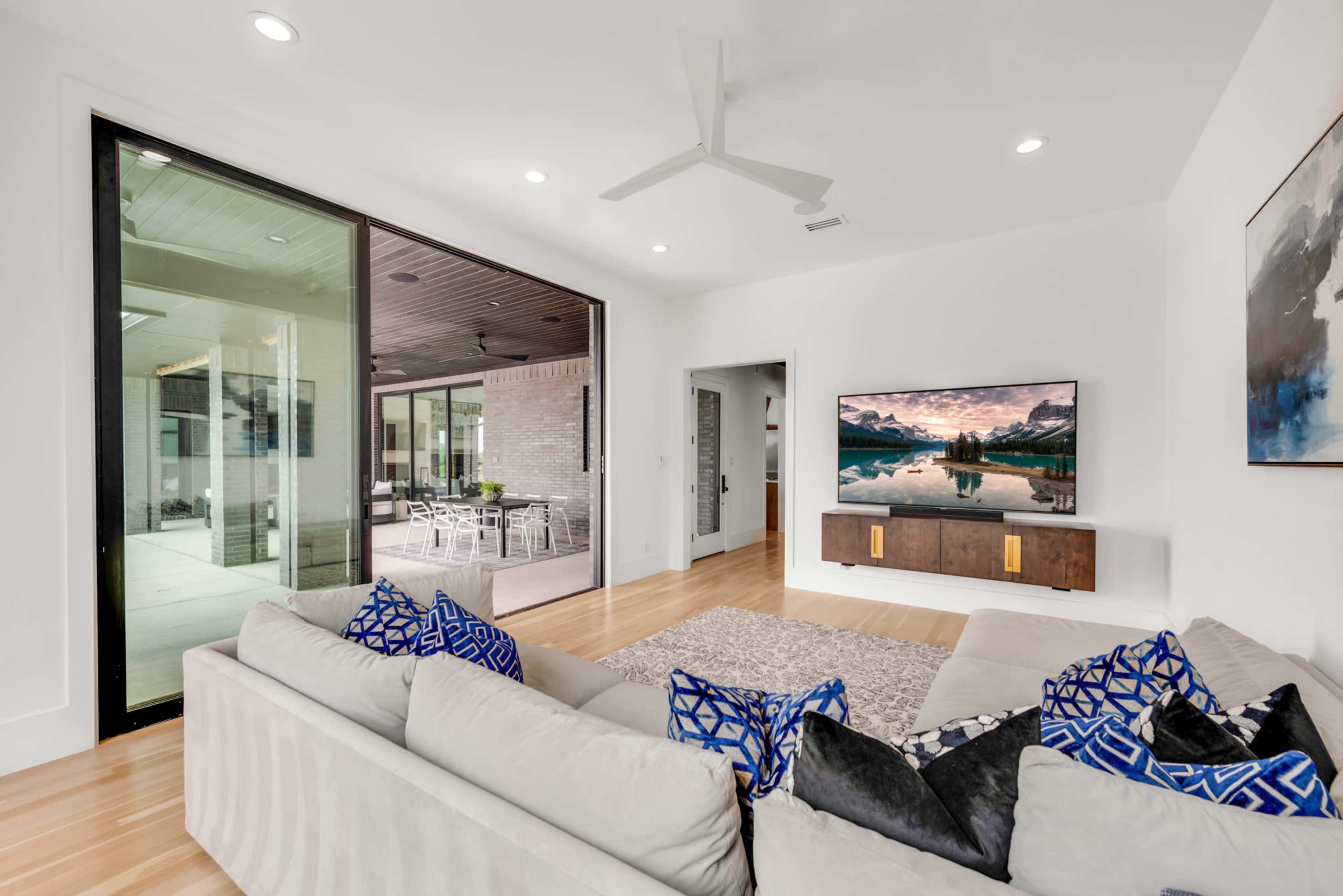 A modern living room features a large sectional sofa with blue and white patterned pillows, a wall-mounted television, and sliding glass doors leading to an outdoor patio.