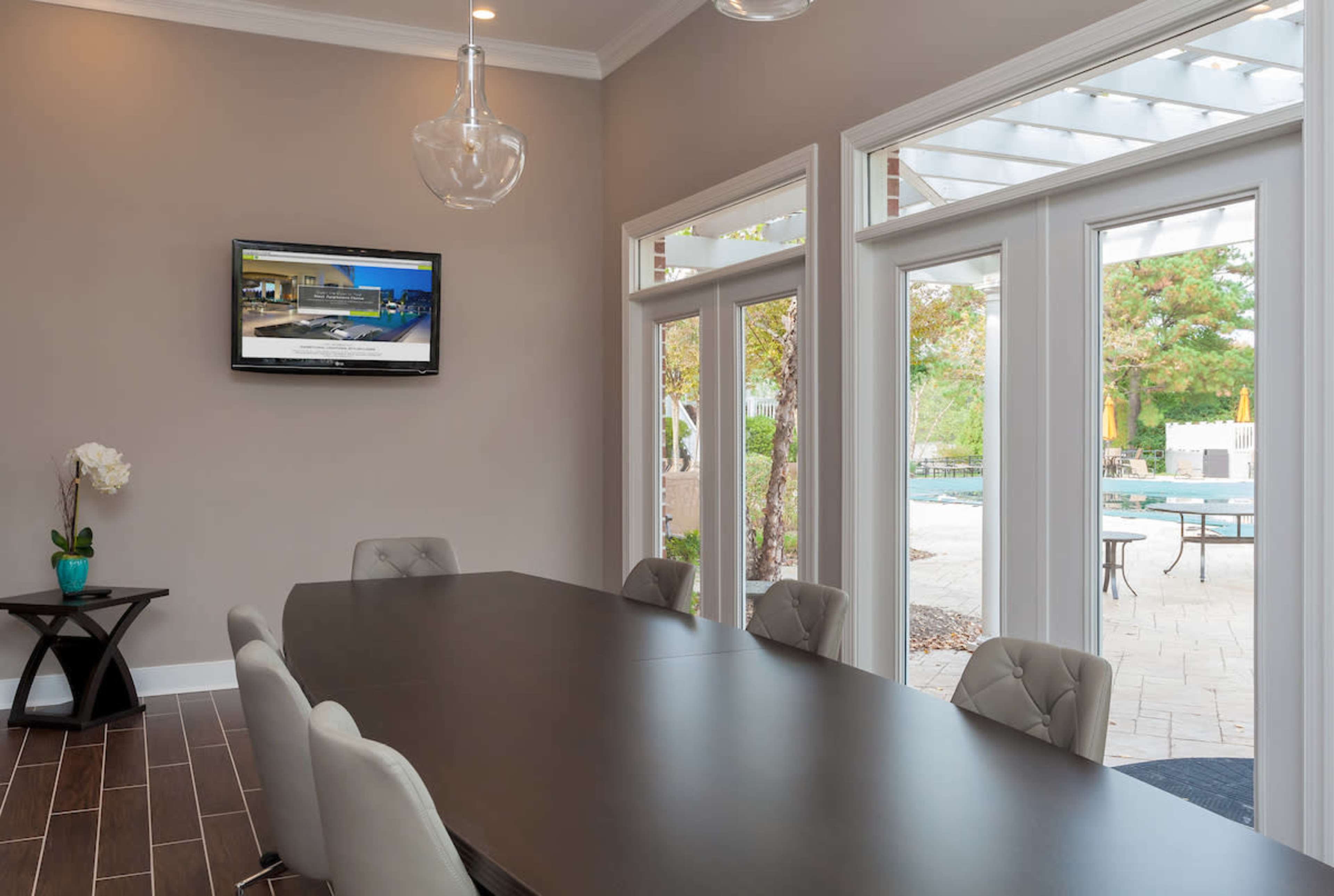 A modern conference room features a long table with upholstered chairs and a television mounted on the wall, with large windows providing a view of an outdoor patio.