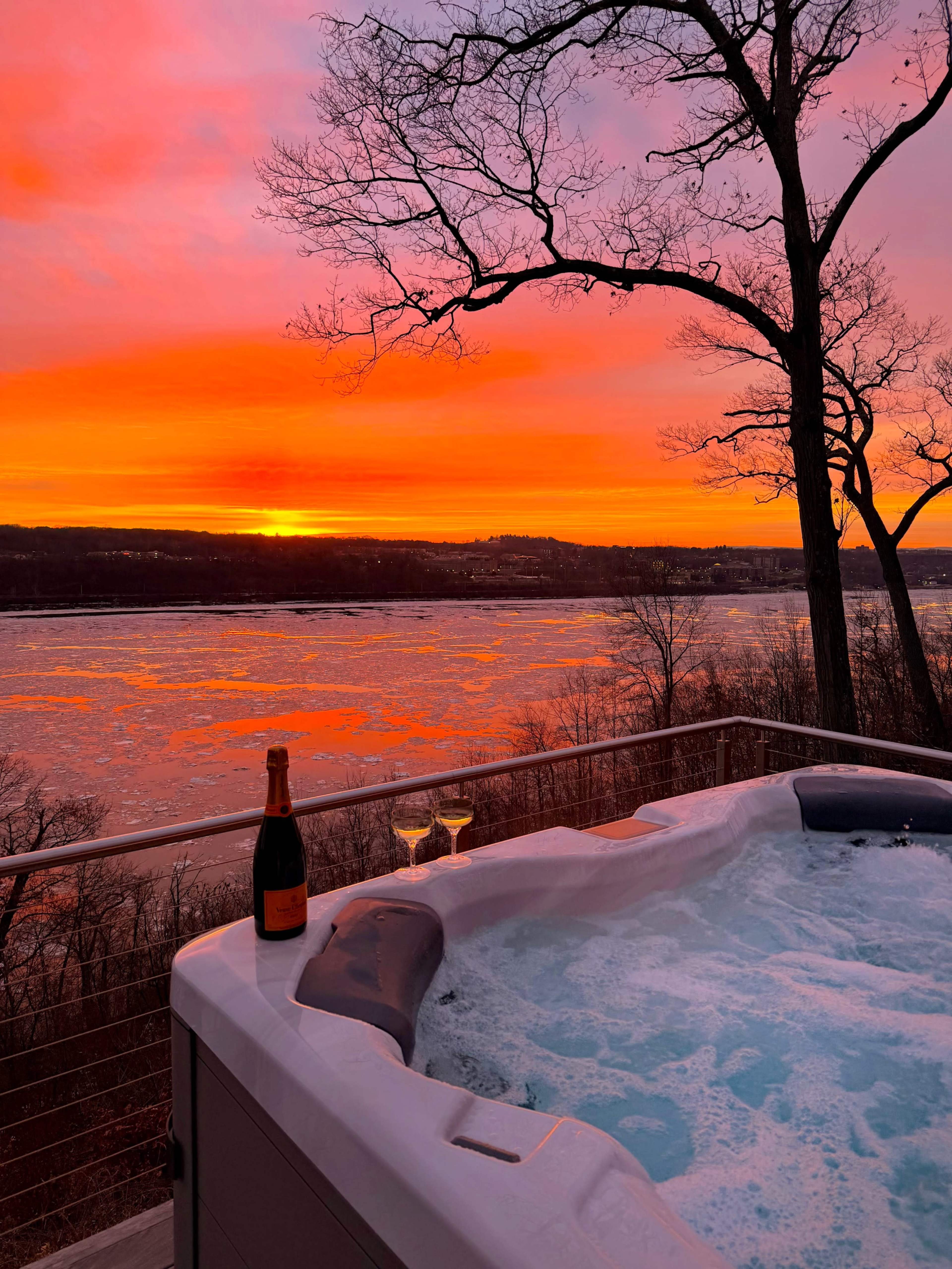 A hot tub overlooking a river at sunset, with a bottle of champagne and two glasses nearby.