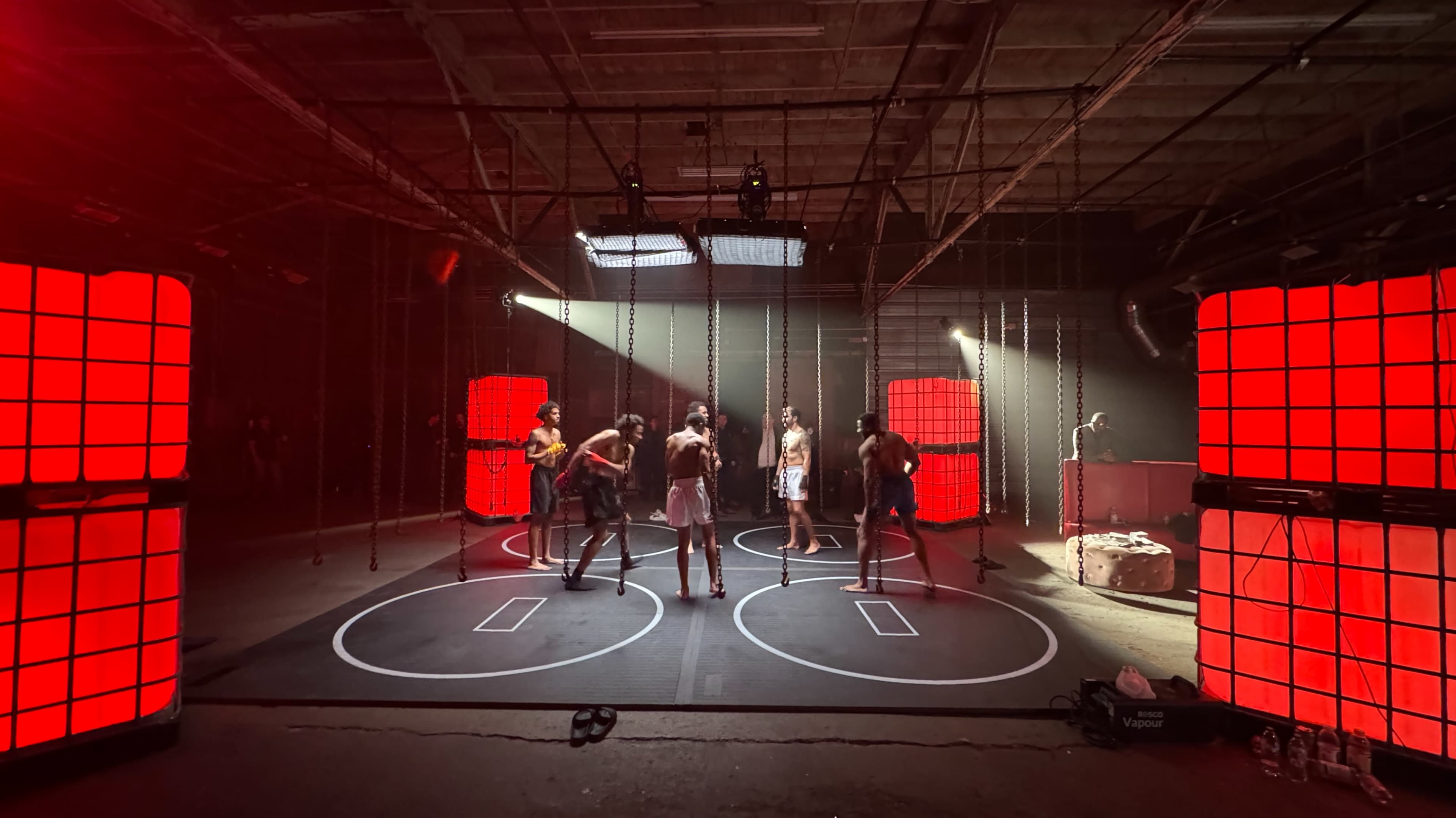 A group of seven people performs on a circular arena surrounded by large red light panels inside an industrial space.
