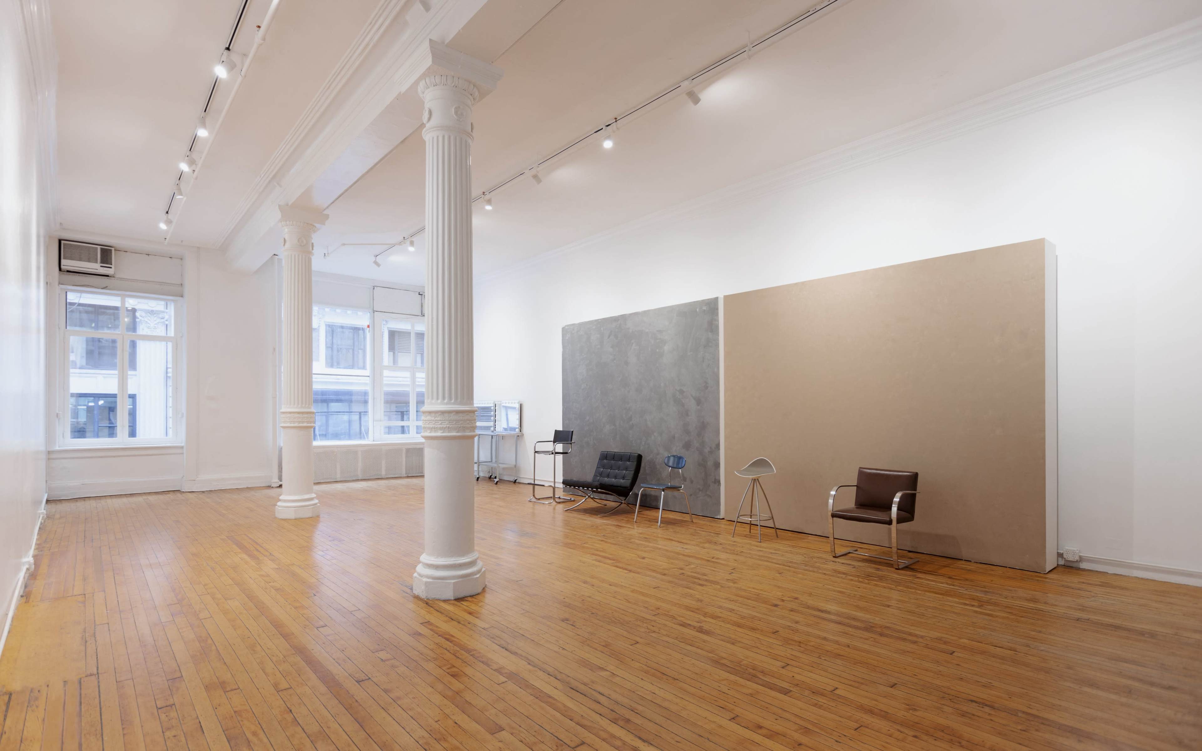 The image shows a spacious, empty room with wooden flooring, tall white columns, and a minimalist design featuring two chairs and a large wall panel.