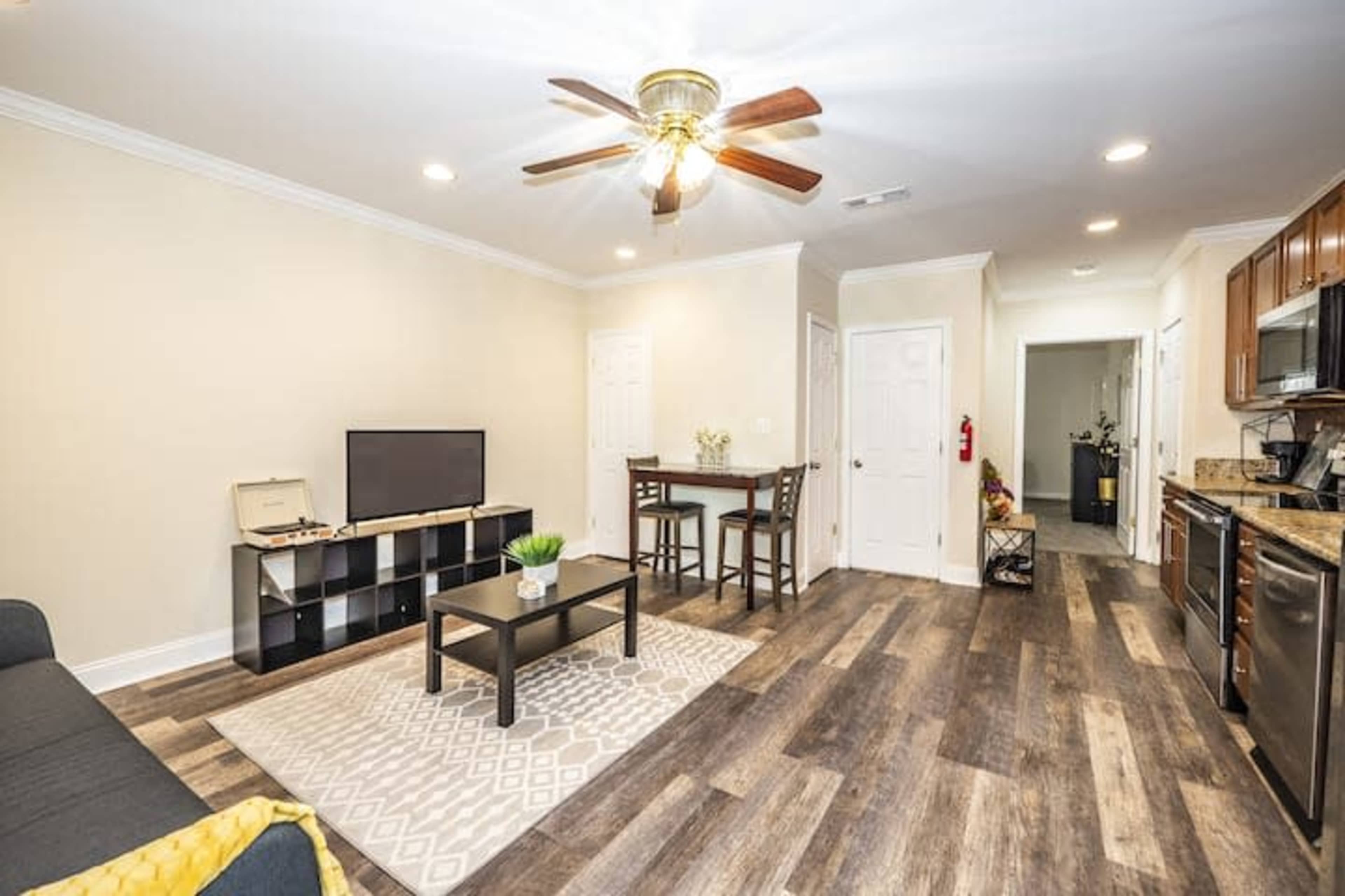A modern living space with a television, a small dining table, a dark sofa, and a kitchen area featuring wooden cabinets and stainless steel appliances.