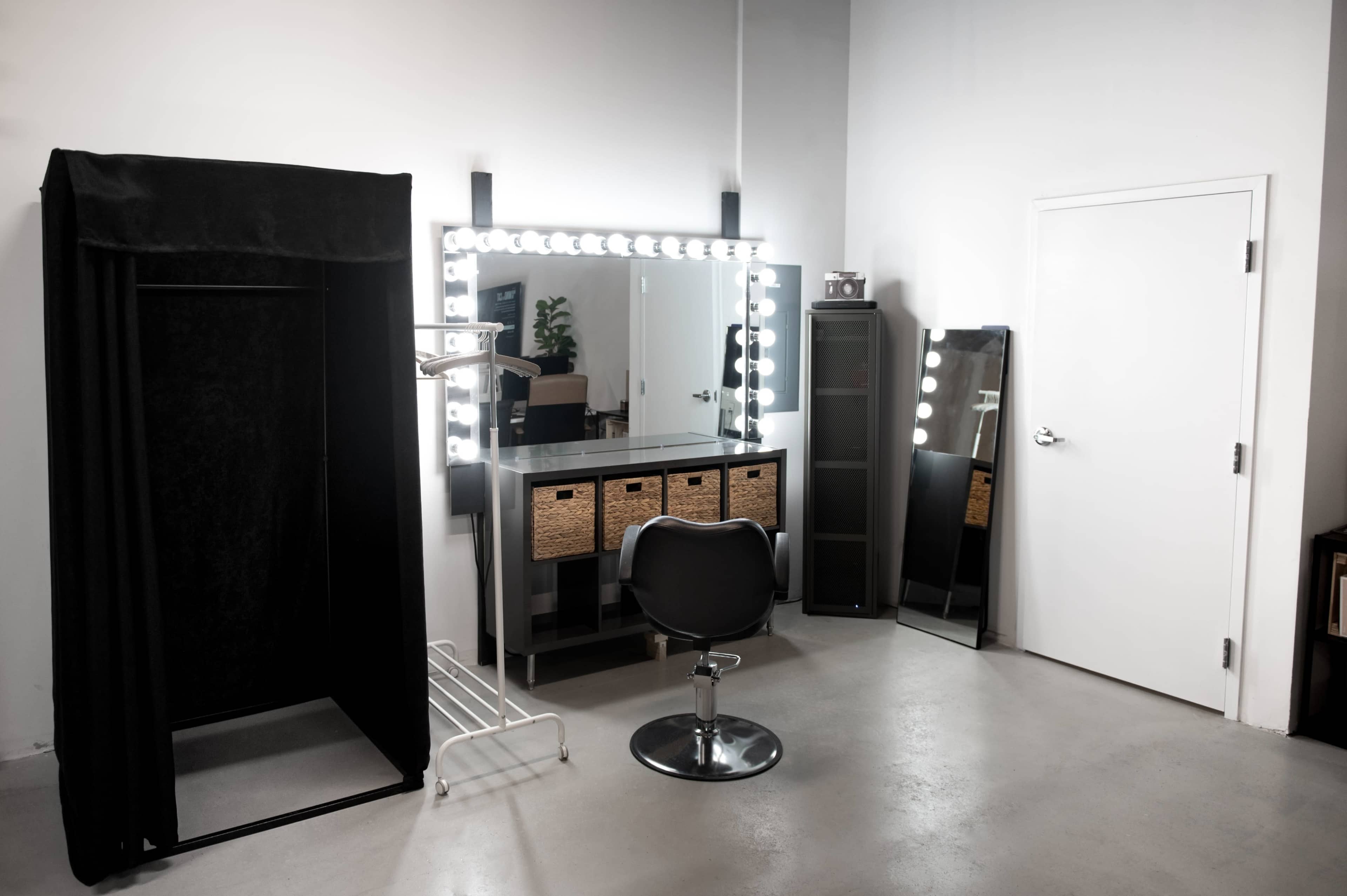 A well-lit vanity area features a large mirror with illuminated bulbs, a black dressing area, a chair, and storage drawers against a plain wall.