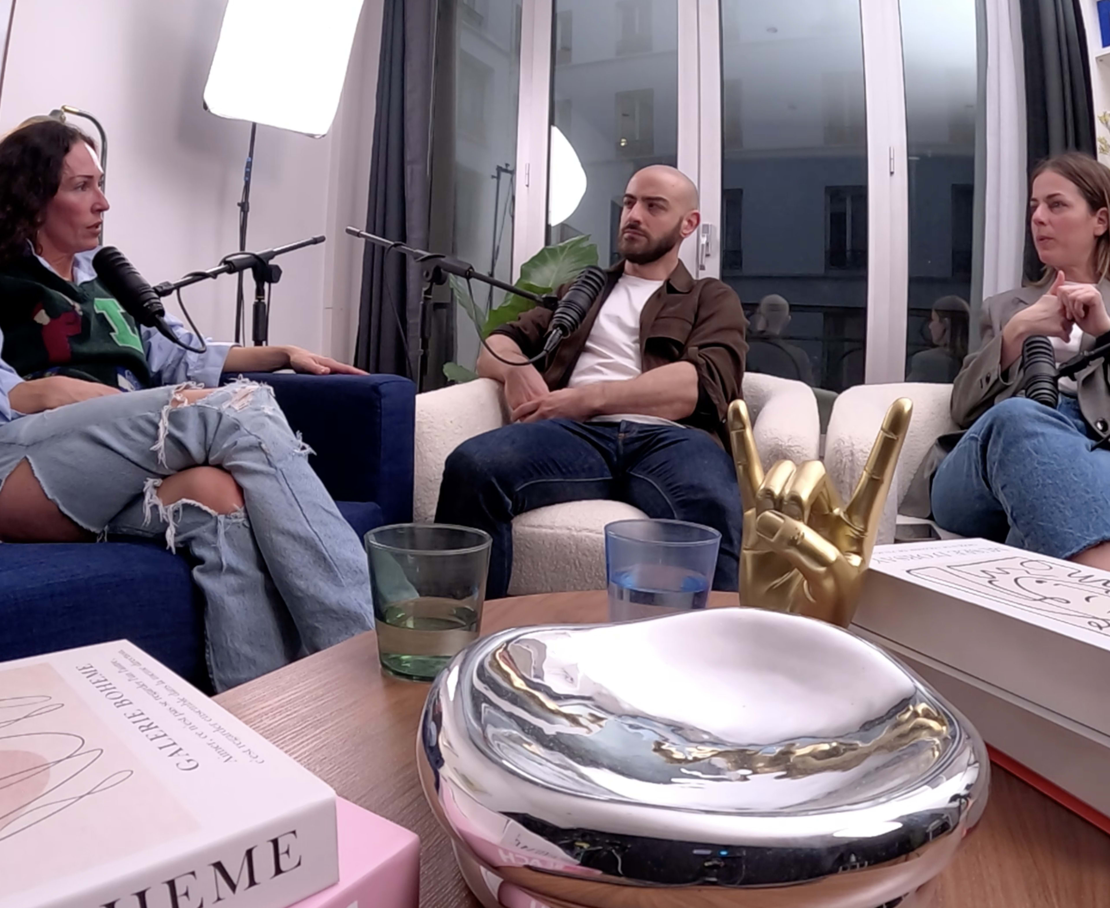 Three people are sitting in a well-lit room engaged in a discussion, with microphones in front of them and various decorative items on the table.