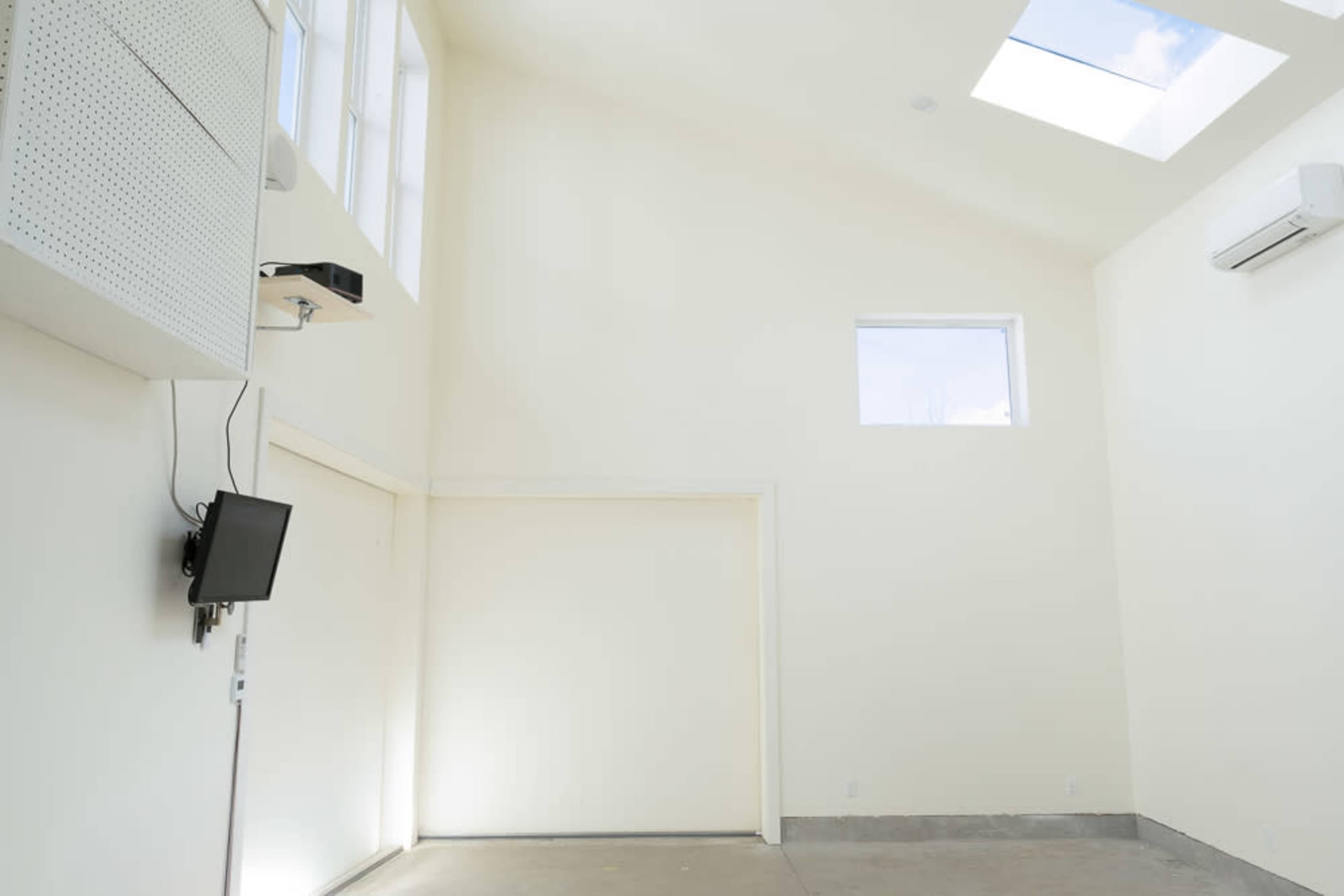 The image shows an empty room with high ceilings, white walls, a ceiling skylight, a small window, and a wall-mounted television.