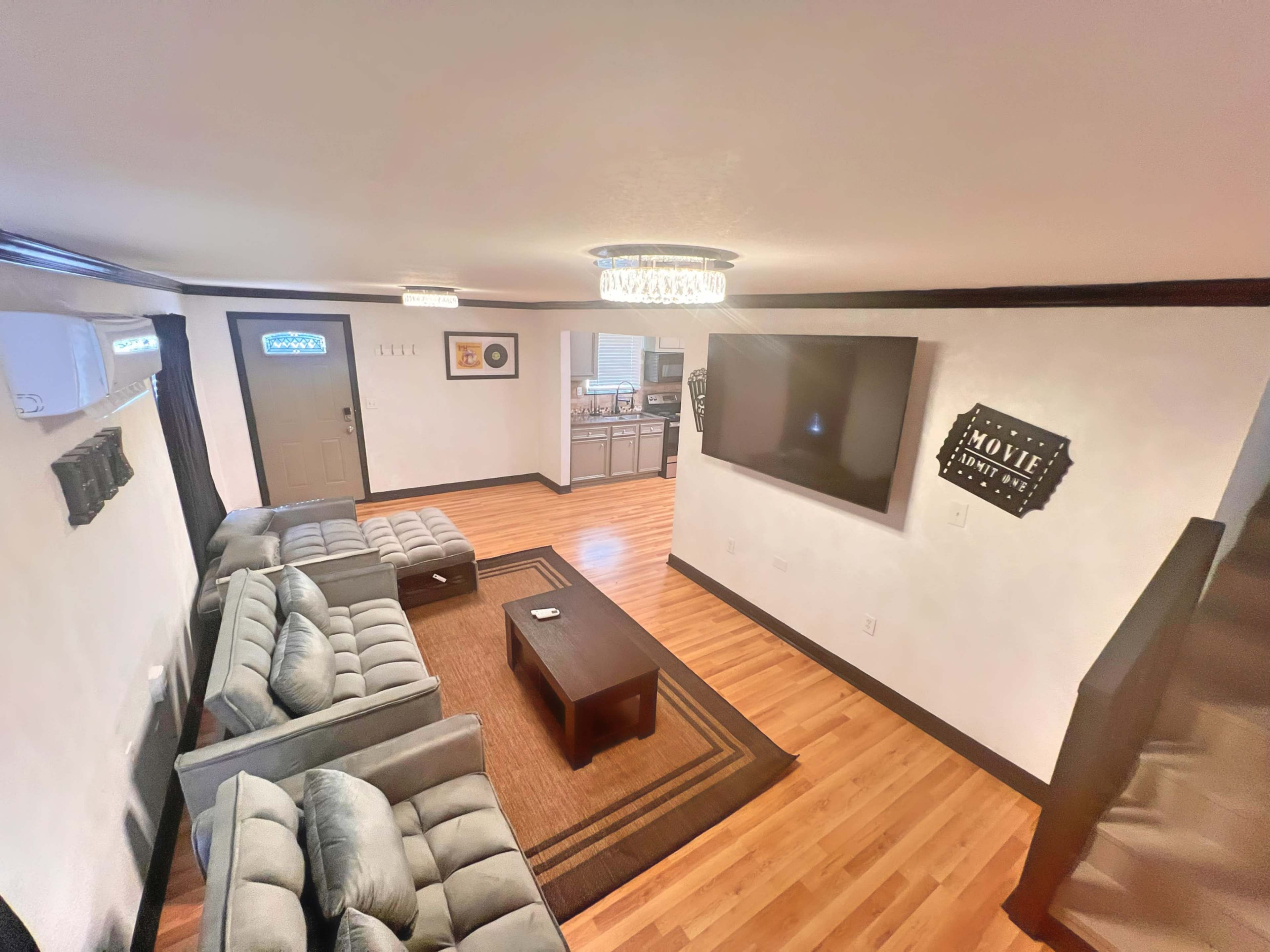 The image shows a living room with gray sofas, a coffee table, a TV mounted on a wall, and a doorway leading to a kitchen area.