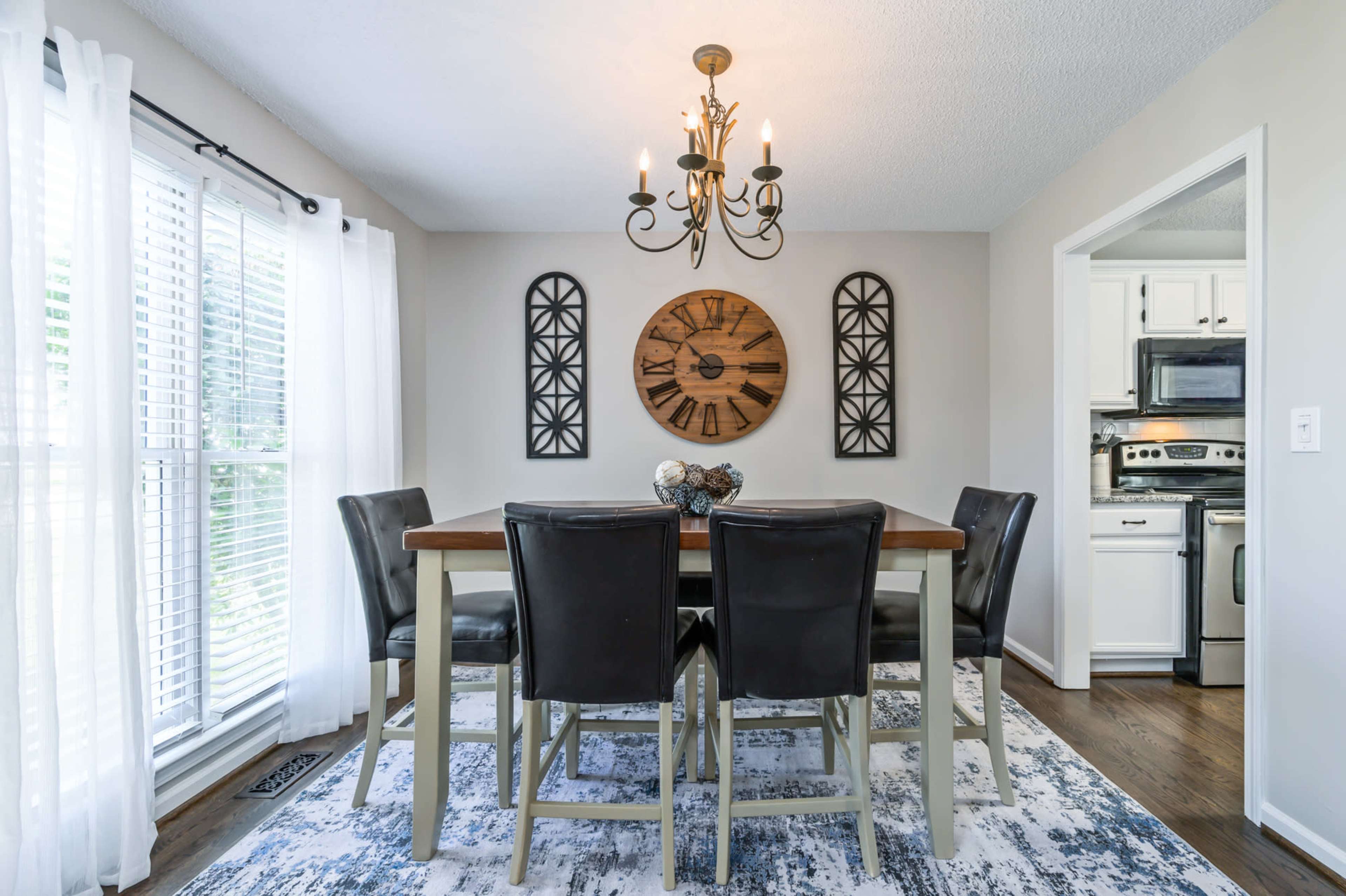 A dining area features a wooden table surrounded by black chairs, with a large clock on the wall and a view of the kitchen in the background.