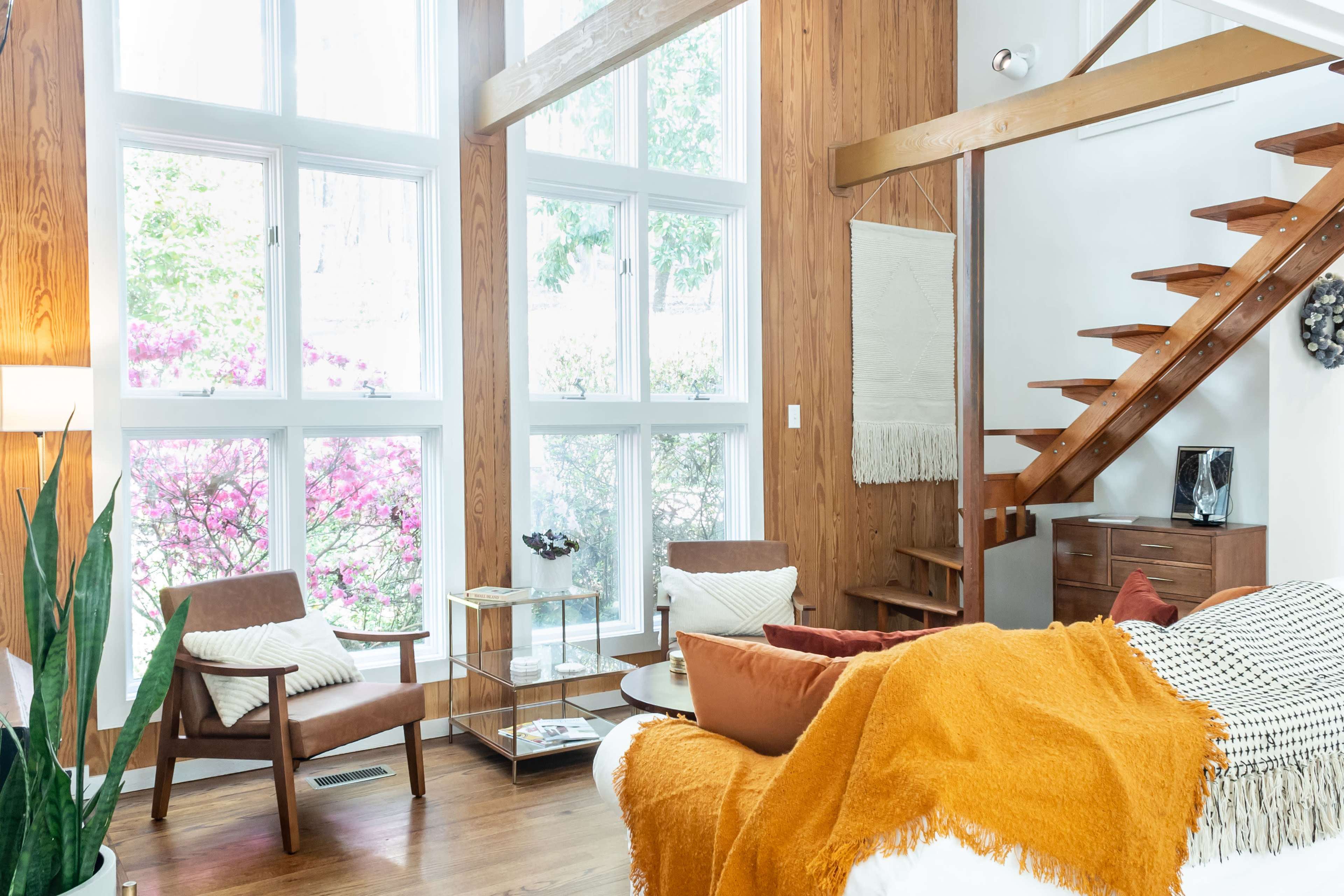 A modern living room with wood-paneled walls, large windows, and a staircase leading to an upper level, featuring comfortable seating and decorative plants.