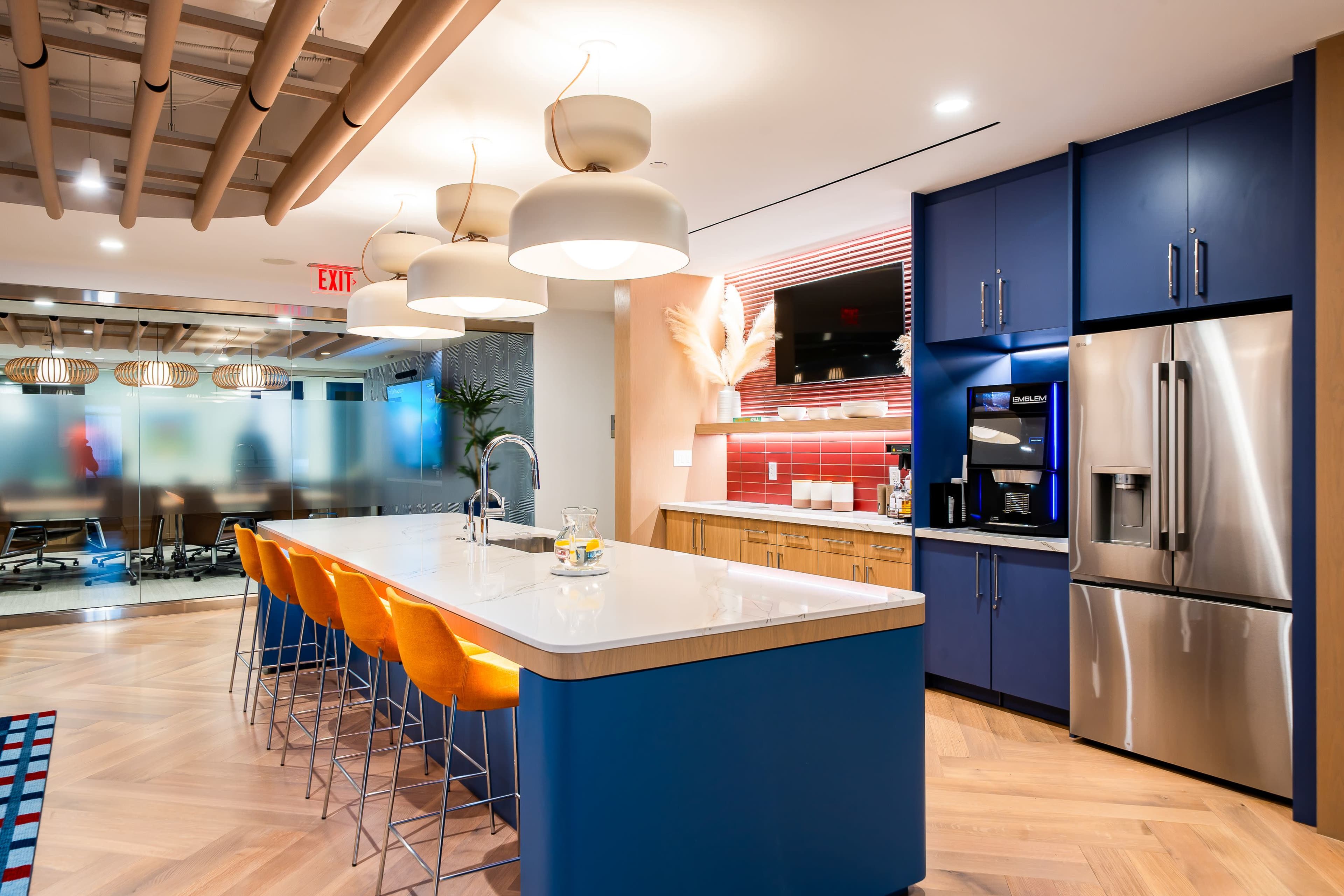 The image shows a modern kitchen with a large island featuring bright orange stools, stainless steel appliances, and a combination of blue and wooden cabinetry.