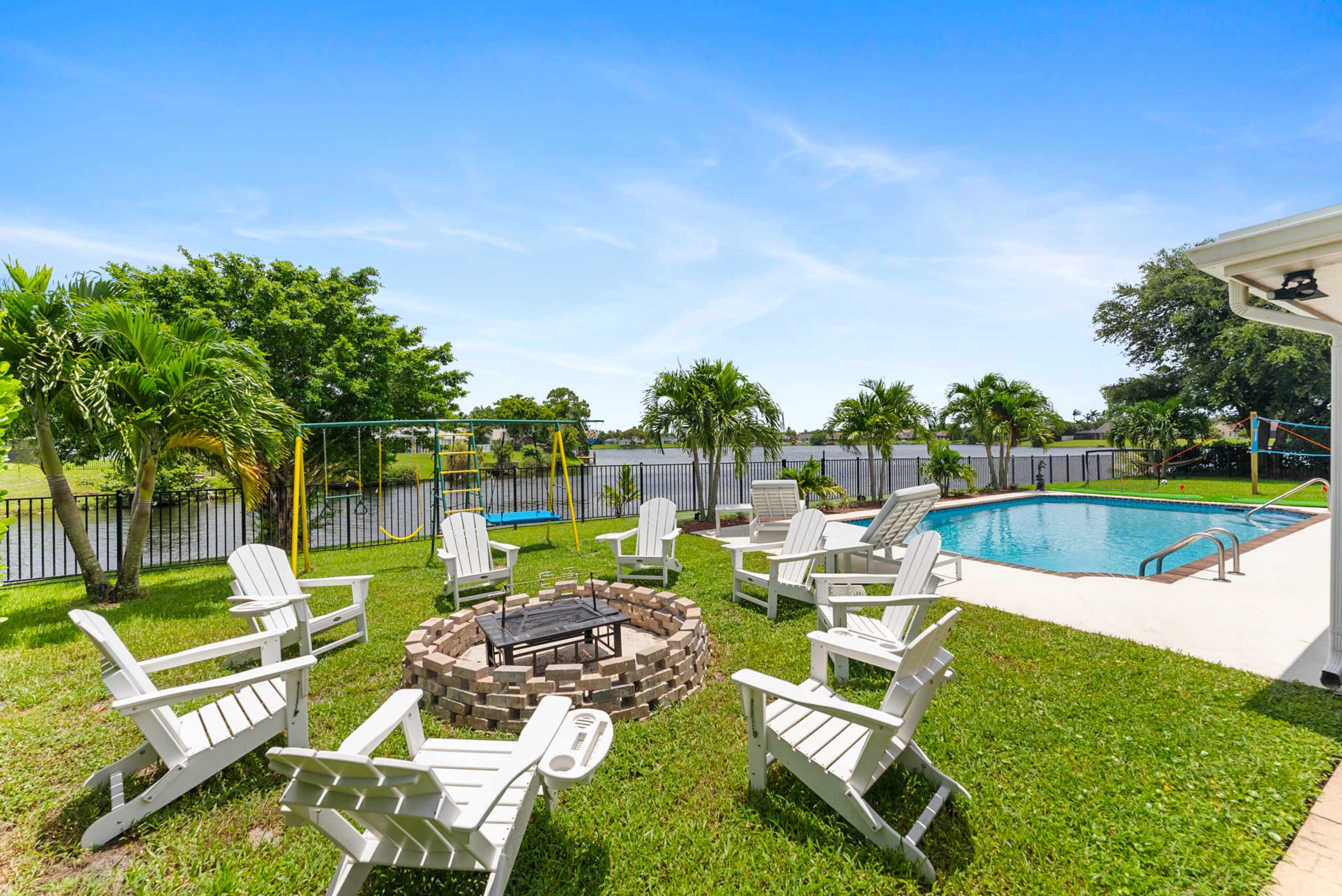 A circular stone fire pit surrounded by white Adirondack chairs is set on the grass next to a swimming pool and a waterfront view.