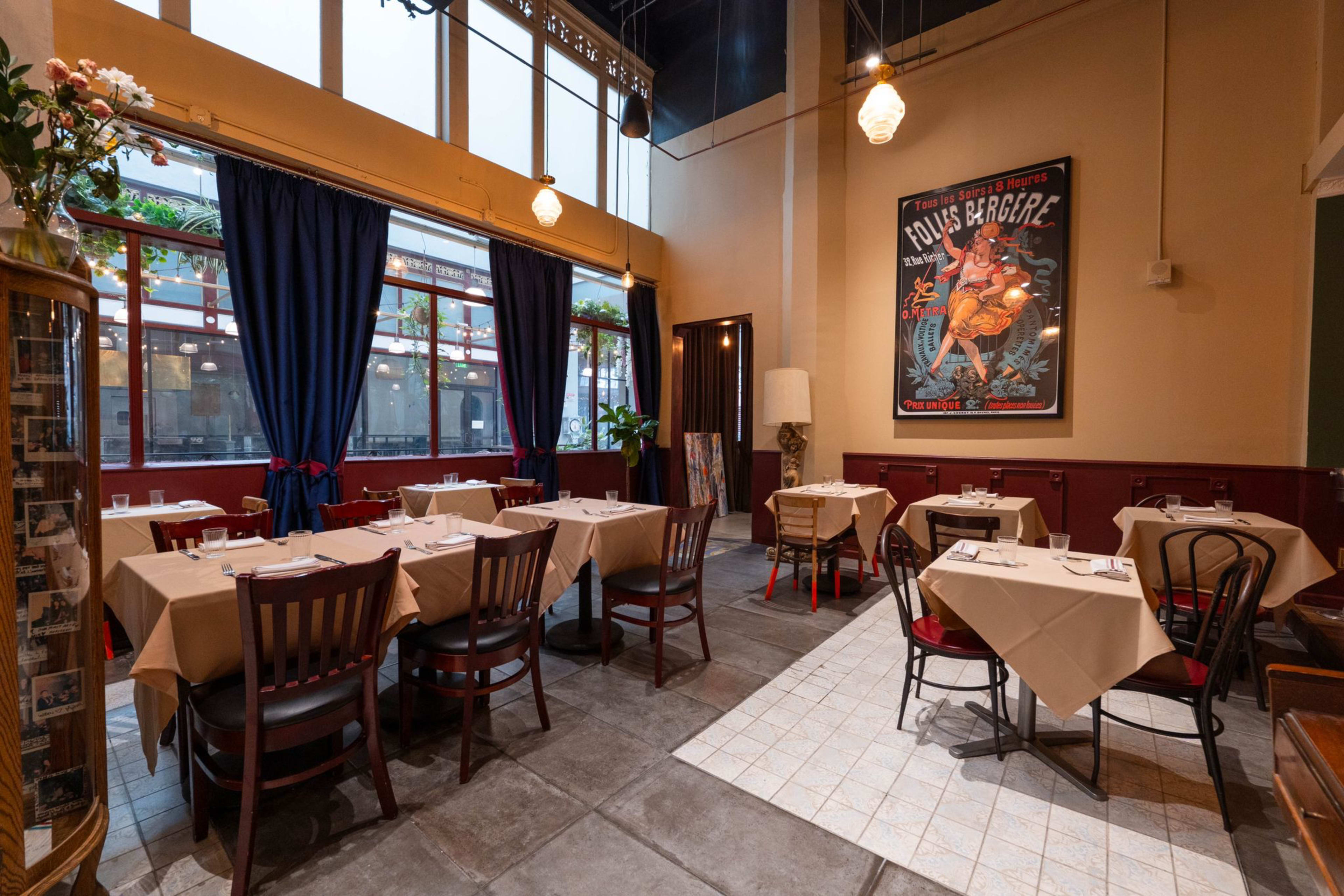 The image shows a restaurant interior featuring several tables with beige tablecloths, large windows with blue curtains, and a colorful poster on the wall.