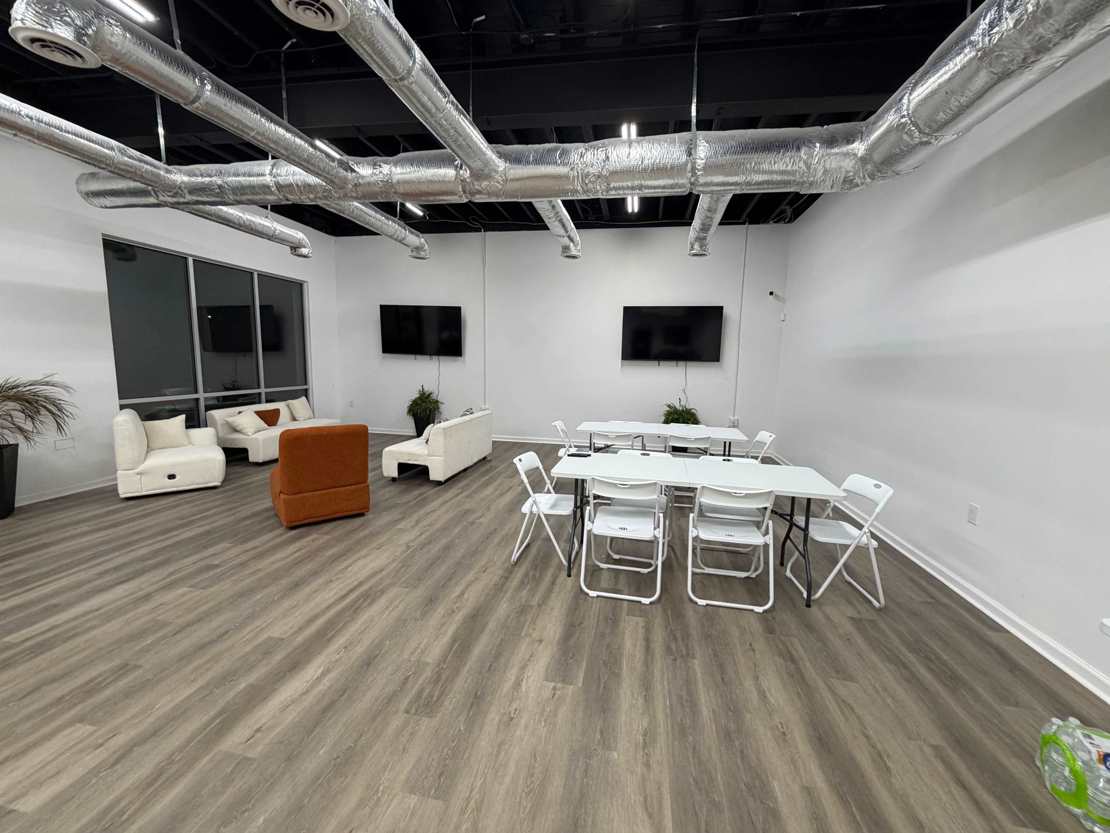 The image shows a modern, minimalist room with wooden flooring, featuring a large table surrounded by folding chairs and a seating area with sofas and plants.