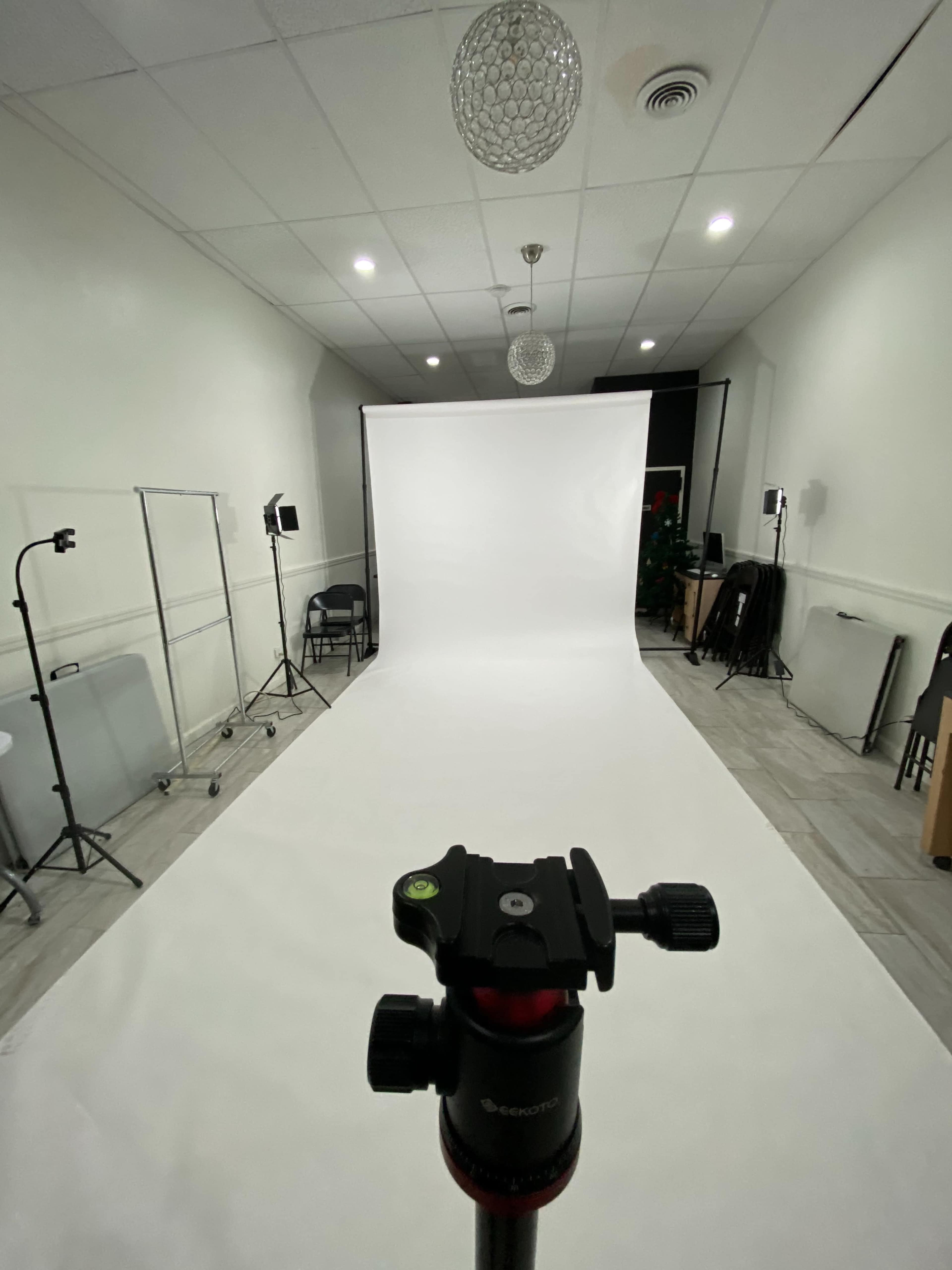 A photography studio is set up with a white backdrop, tripod in the foreground, and various lighting equipment arranged around the room.