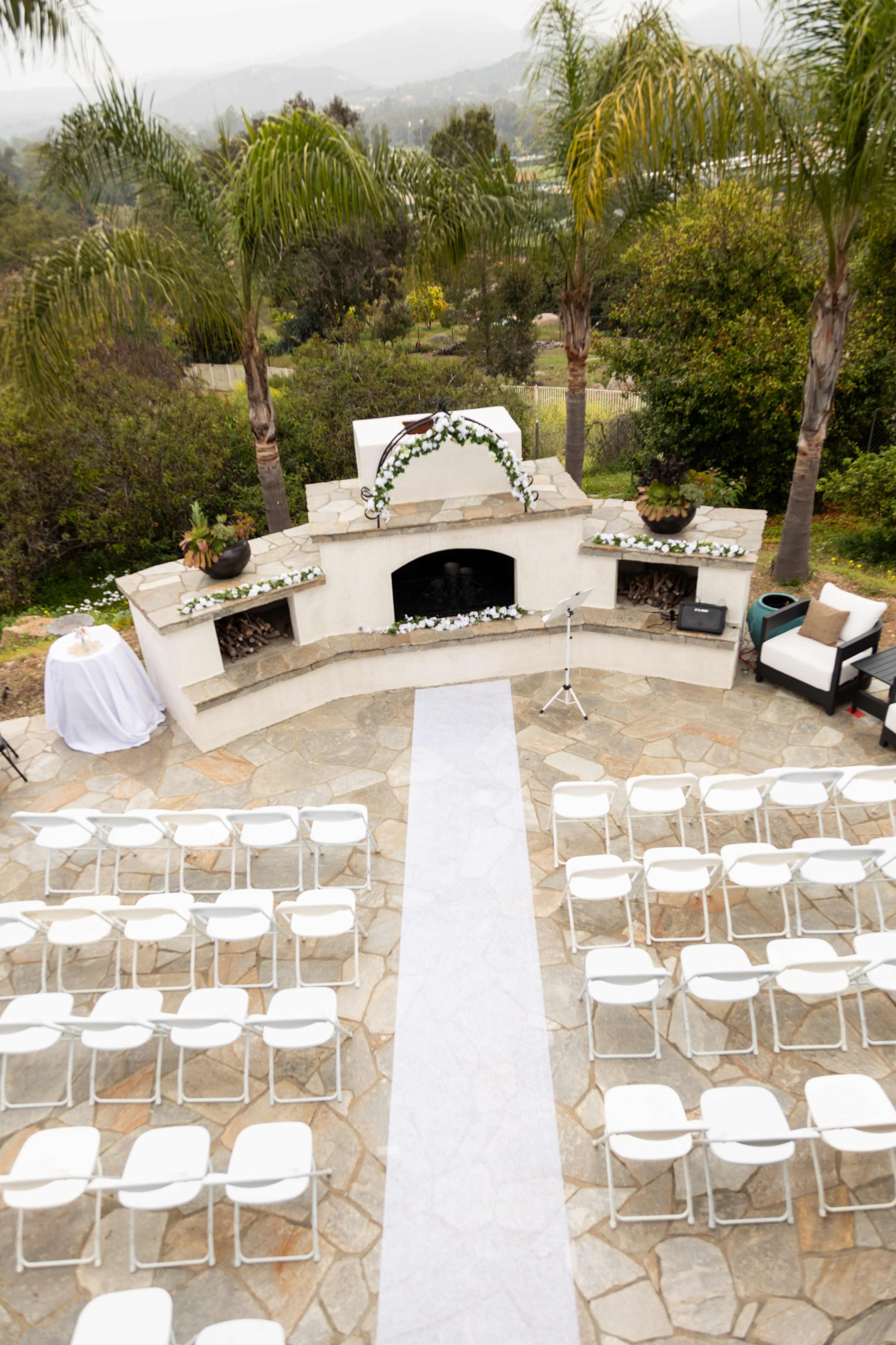An outdoor wedding venue with rows of white chairs arranged in front of a decorated altar and a stone fireplace, surrounded by greenery and palm trees.