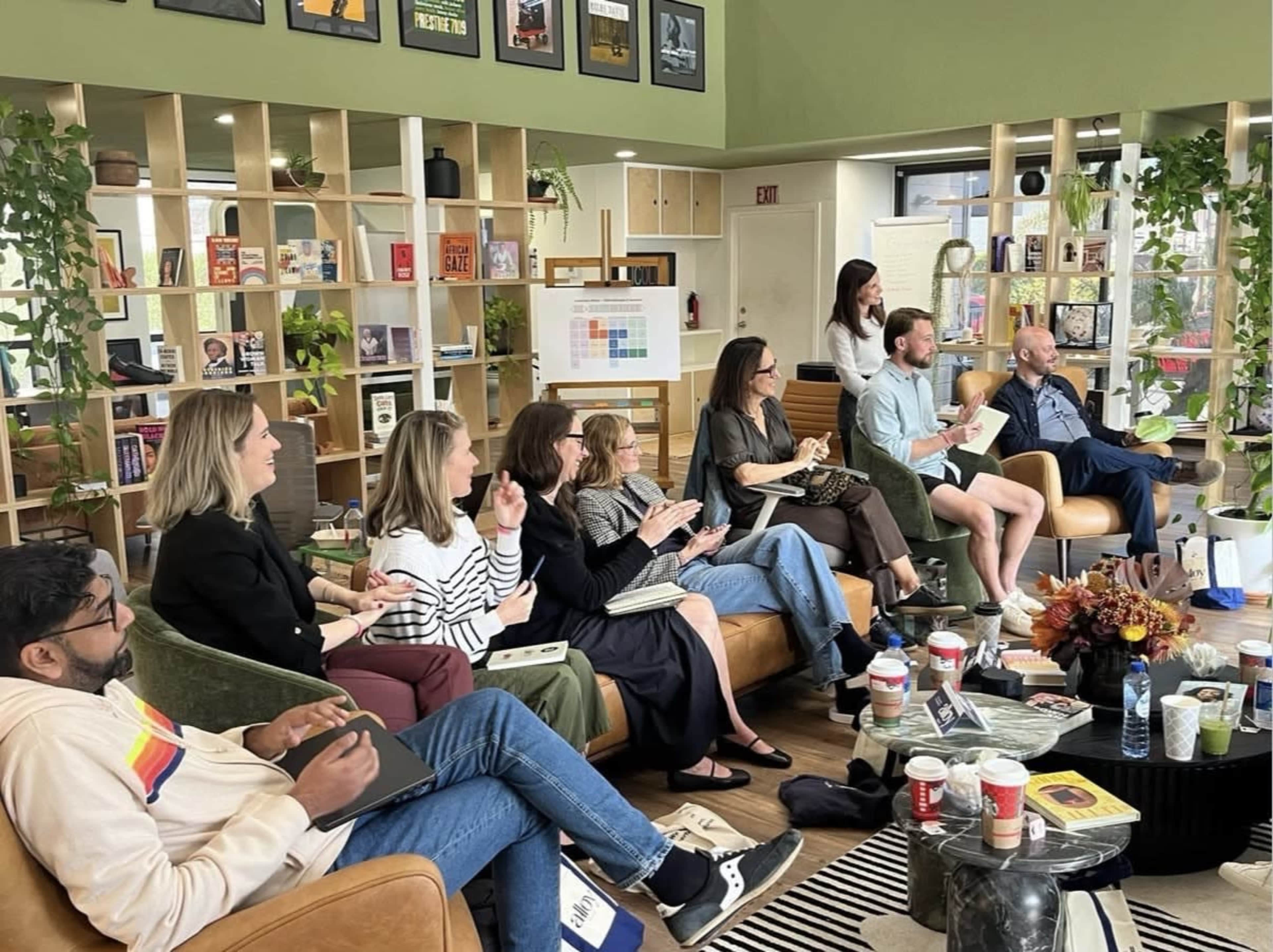 A group of eight people sits in a modern room with bookshelves, engaging in a discussion while holding notebooks and drinks.