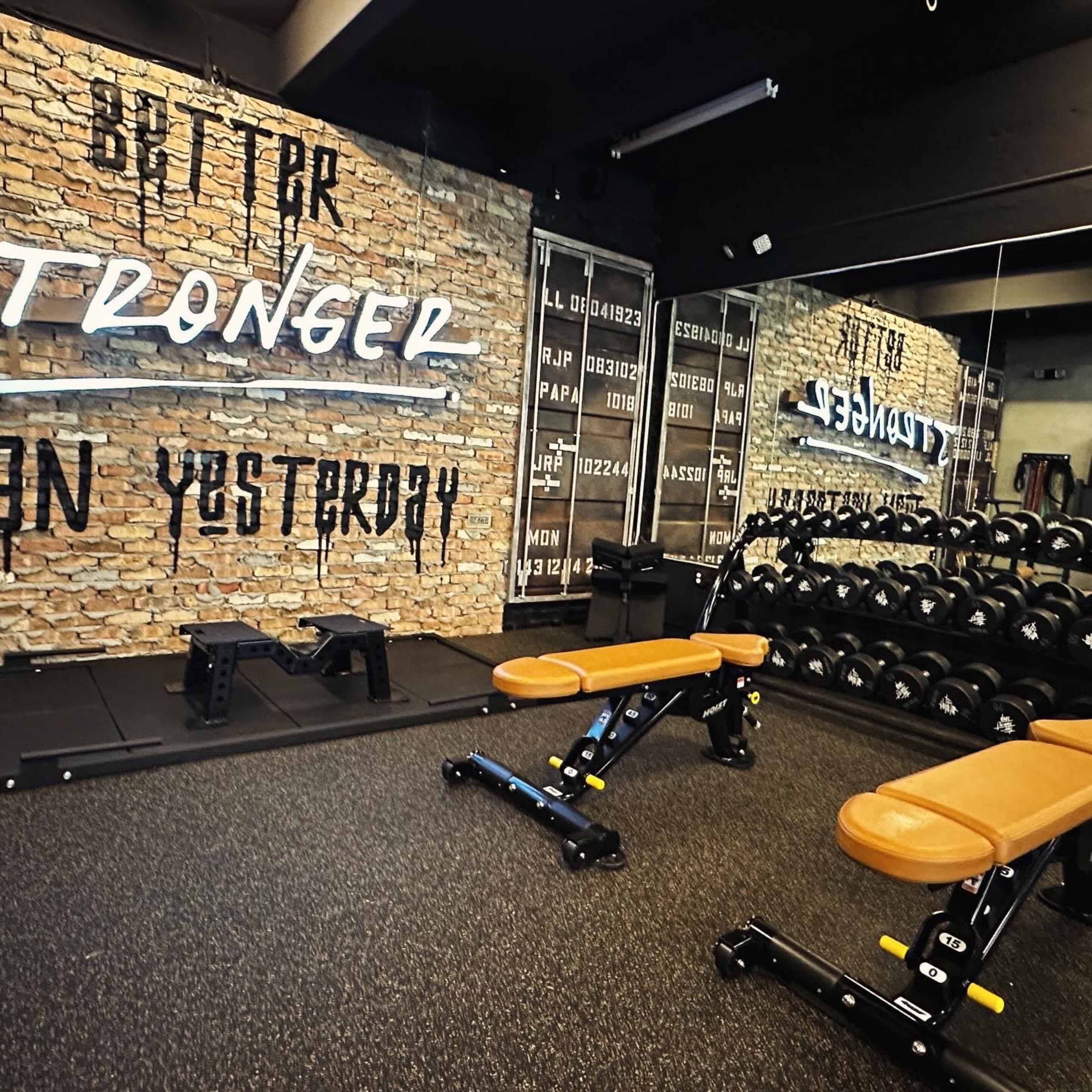 A modern gym interior featuring wooden benches, free weights, and a brick wall with motivational neon signage.