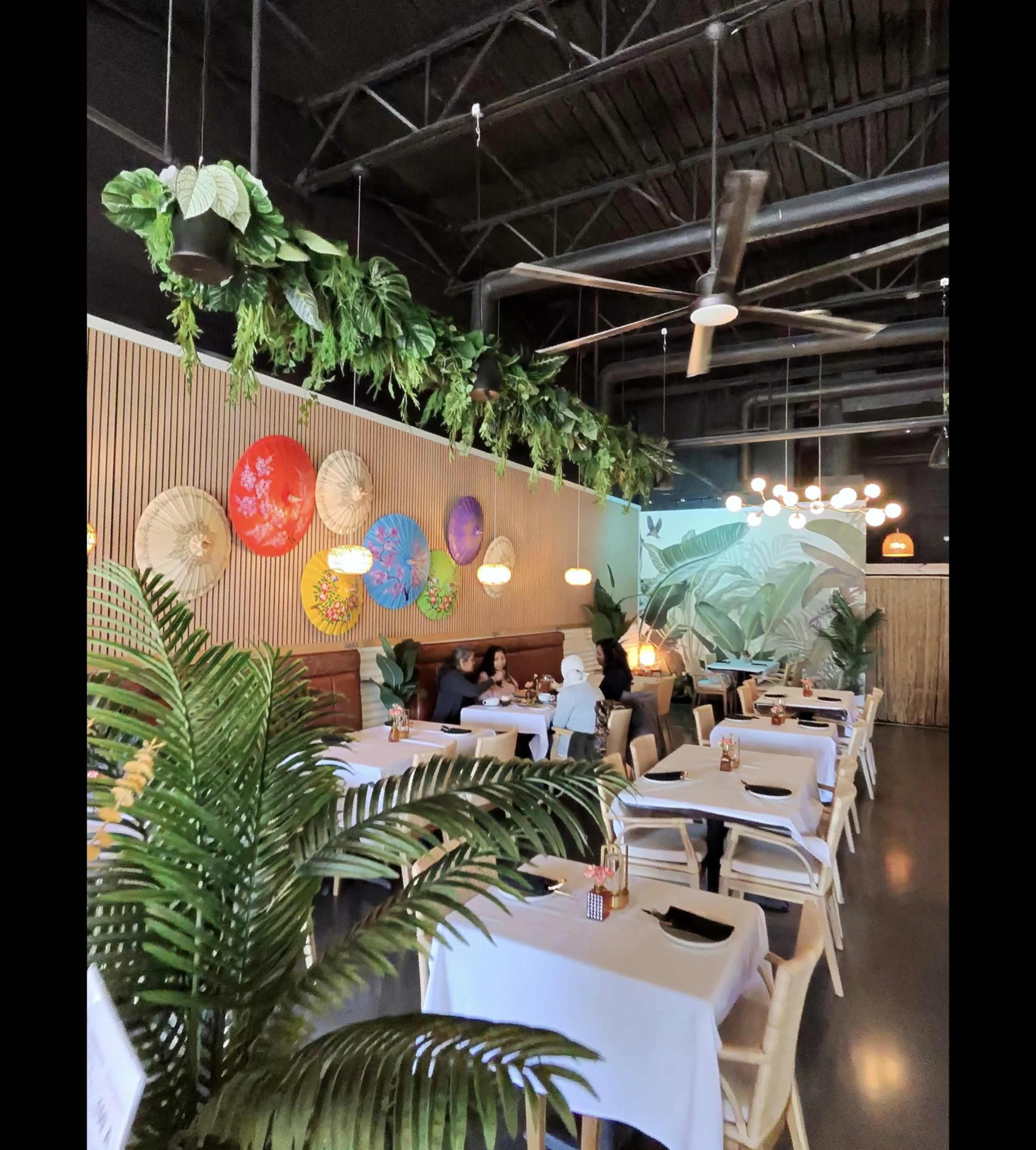 The image shows a modern restaurant interior with white tablecloths, decorative umbrellas on the wall, hanging plants, and a mural of tropical foliage.