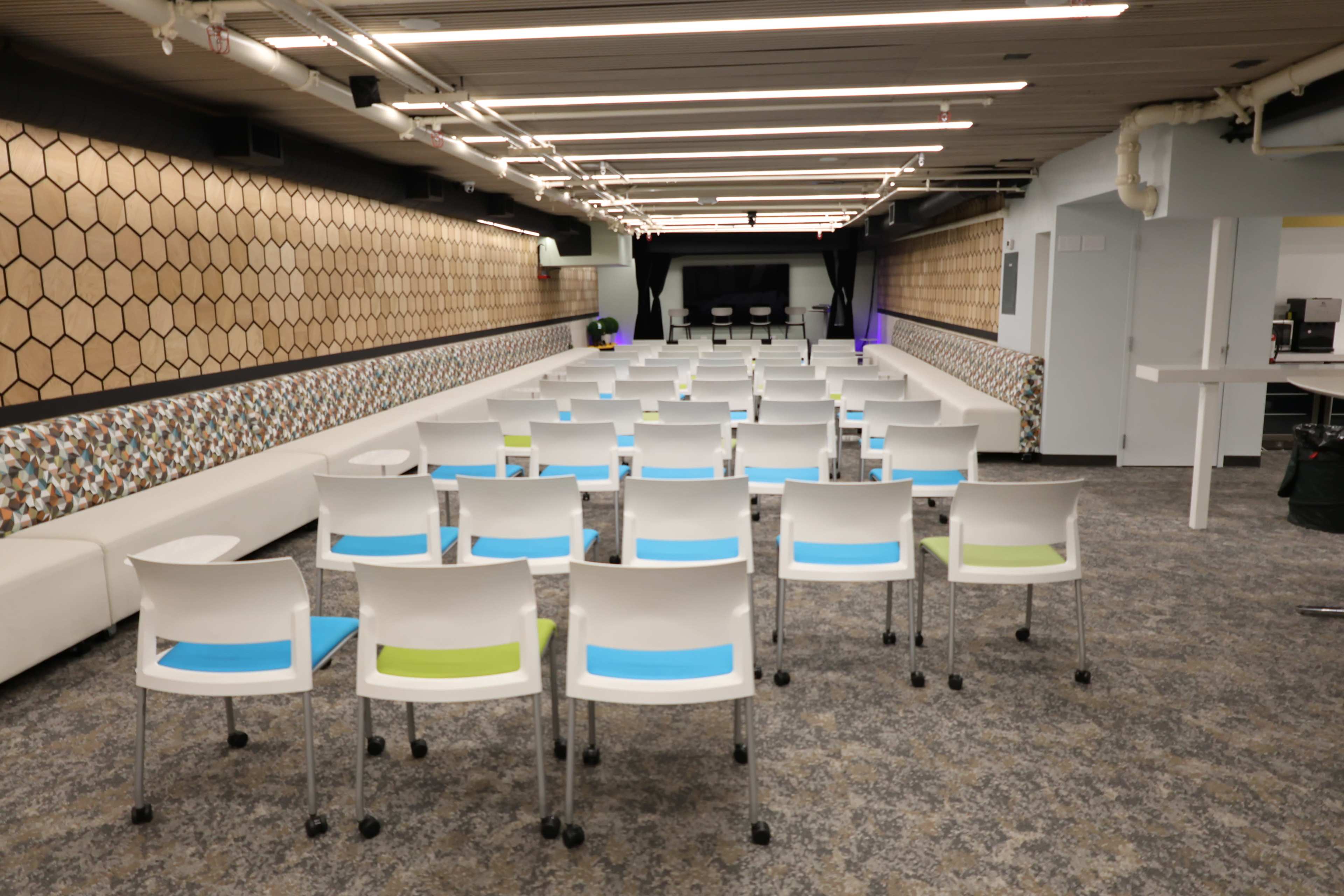 The image shows a spacious auditorium with rows of white chairs, some with colorful seat cushions, facing a stage area at the front.
