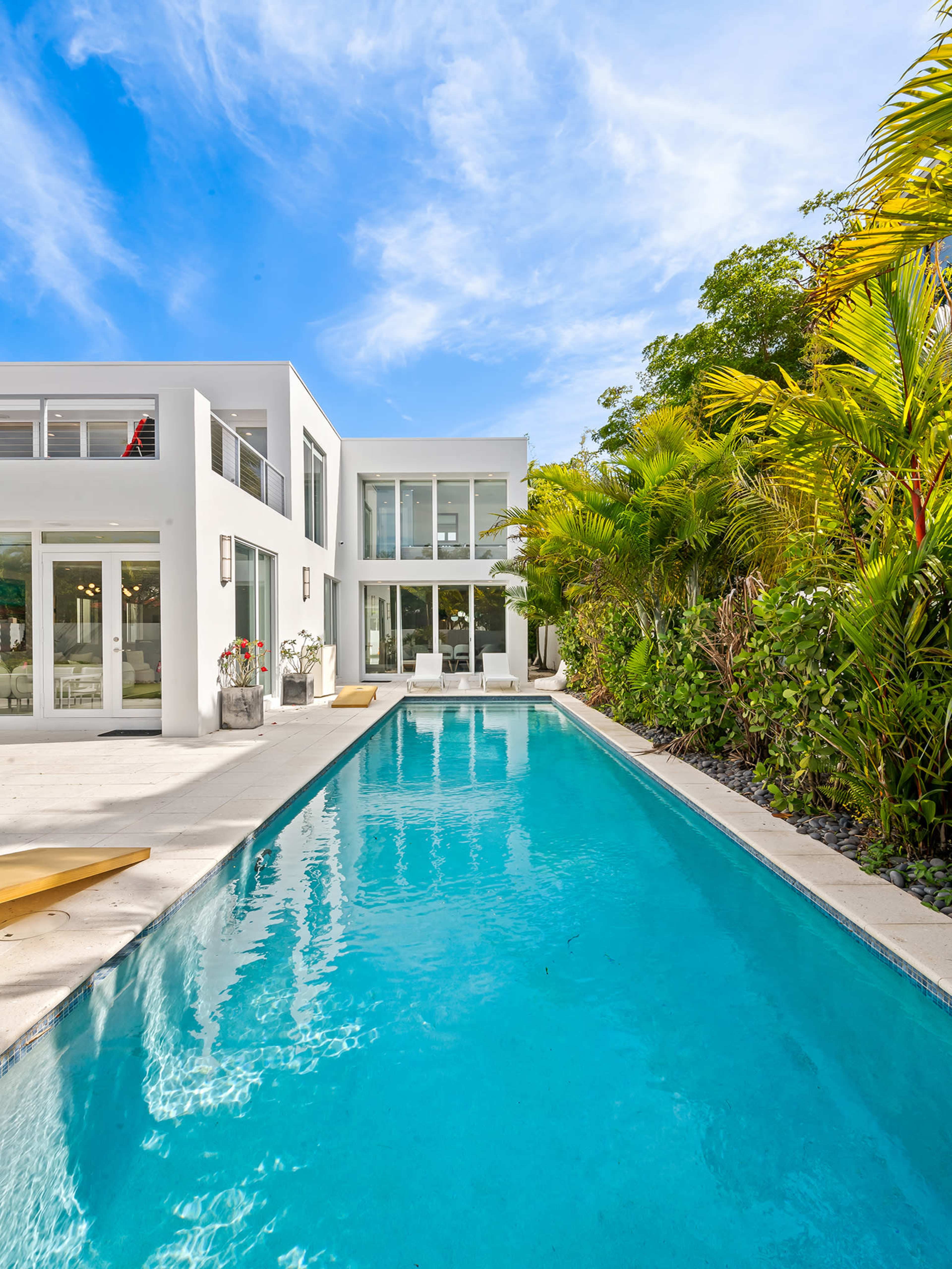 The image shows a modern white house beside a clear swimming pool surrounded by lush greenery and tropical plants.