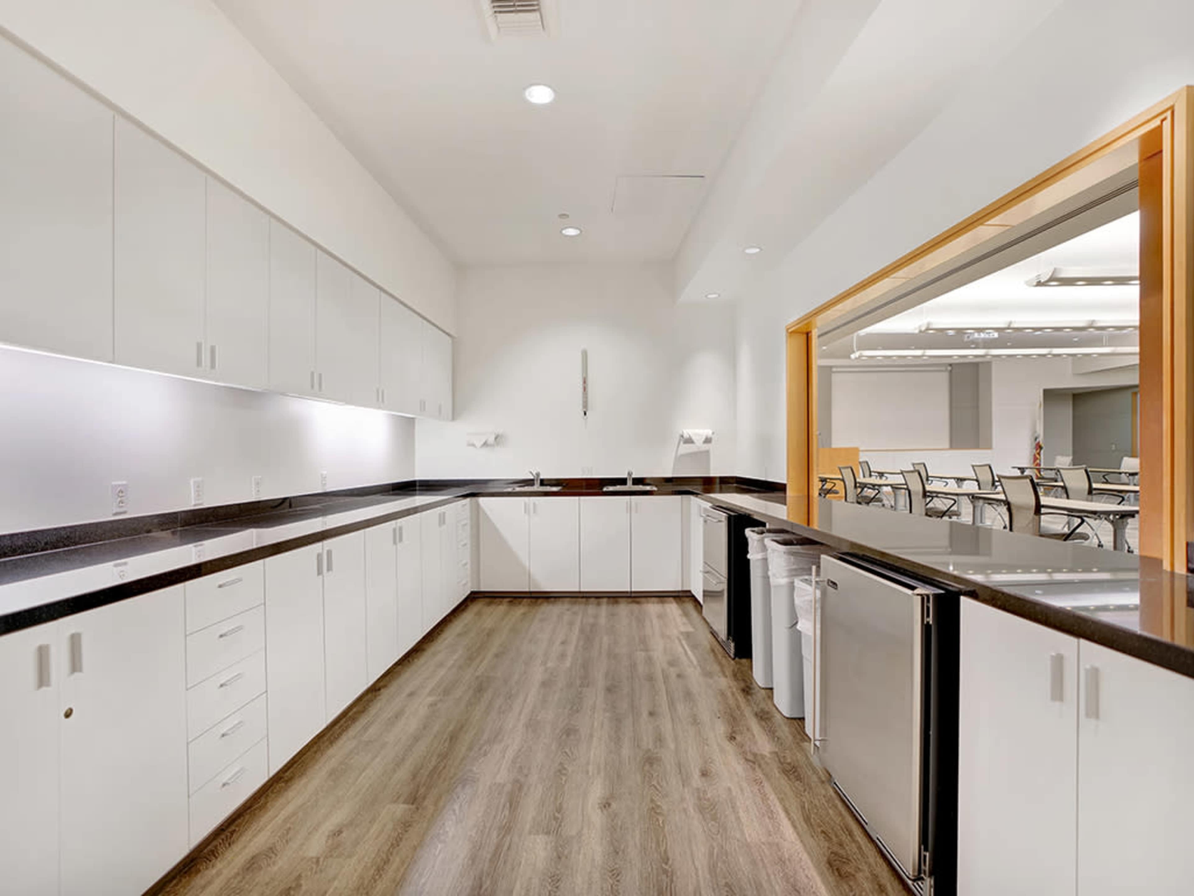 The image shows a spacious kitchen with white cabinetry, a dark countertop, and a polished wooden floor, featuring appliances and a large opening to an adjacent room.