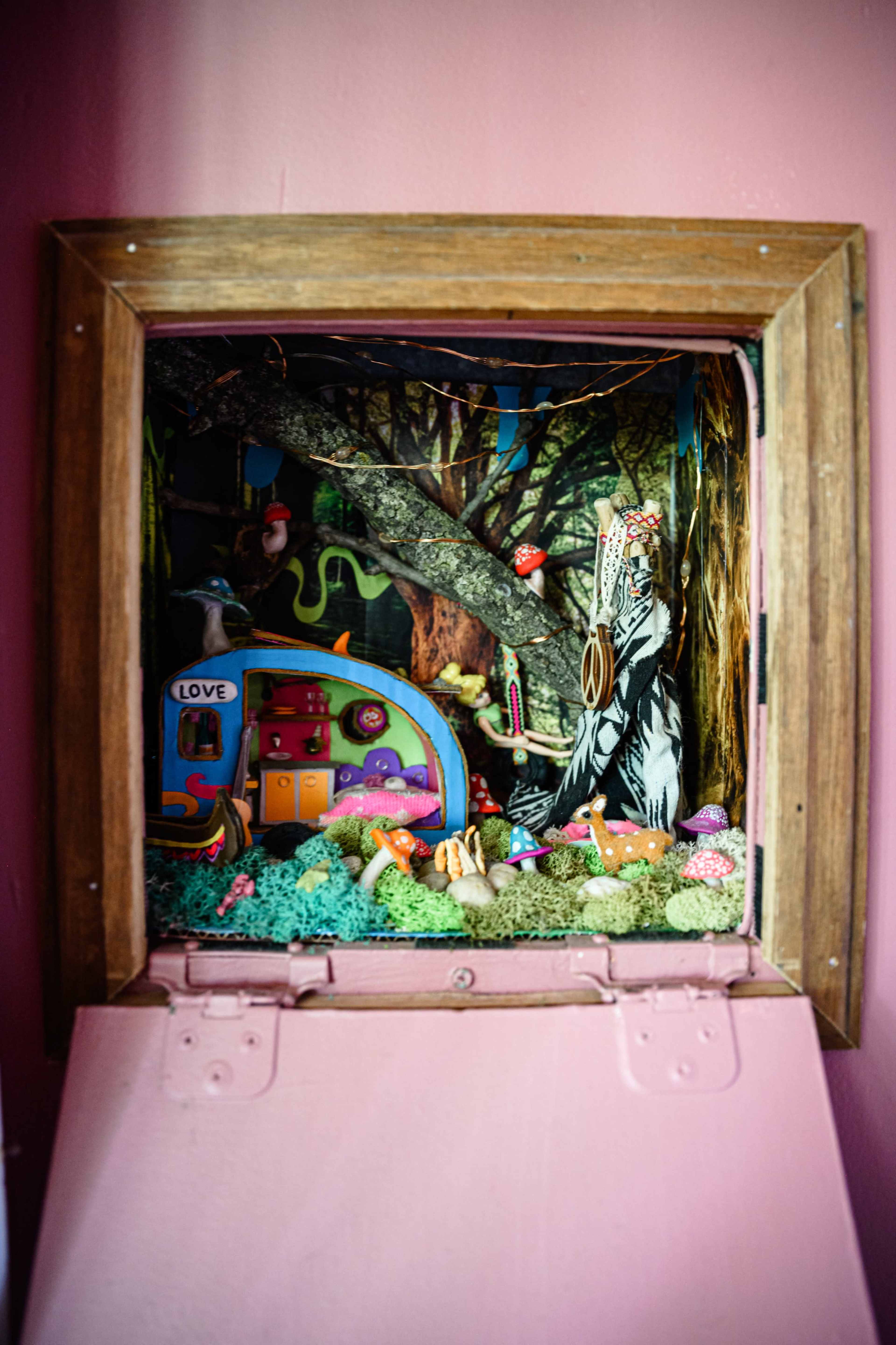 The image shows a miniature diorama inside a wooden frame, featuring a colorful scene with trees, animals, and a small house.