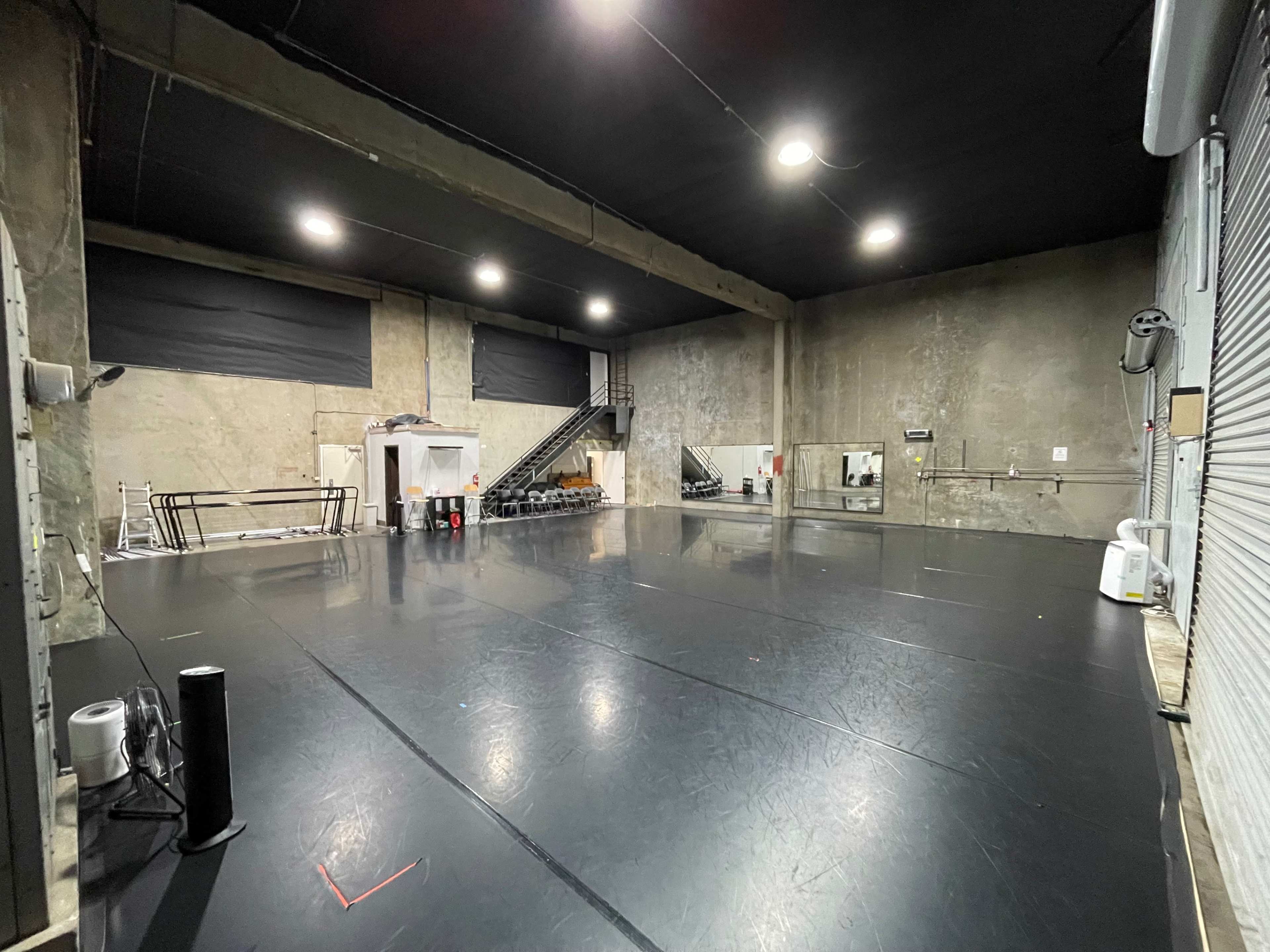 The image shows a large, empty rehearsal space with a black dance floor and concrete walls, featuring a staircase and equipment in the background.