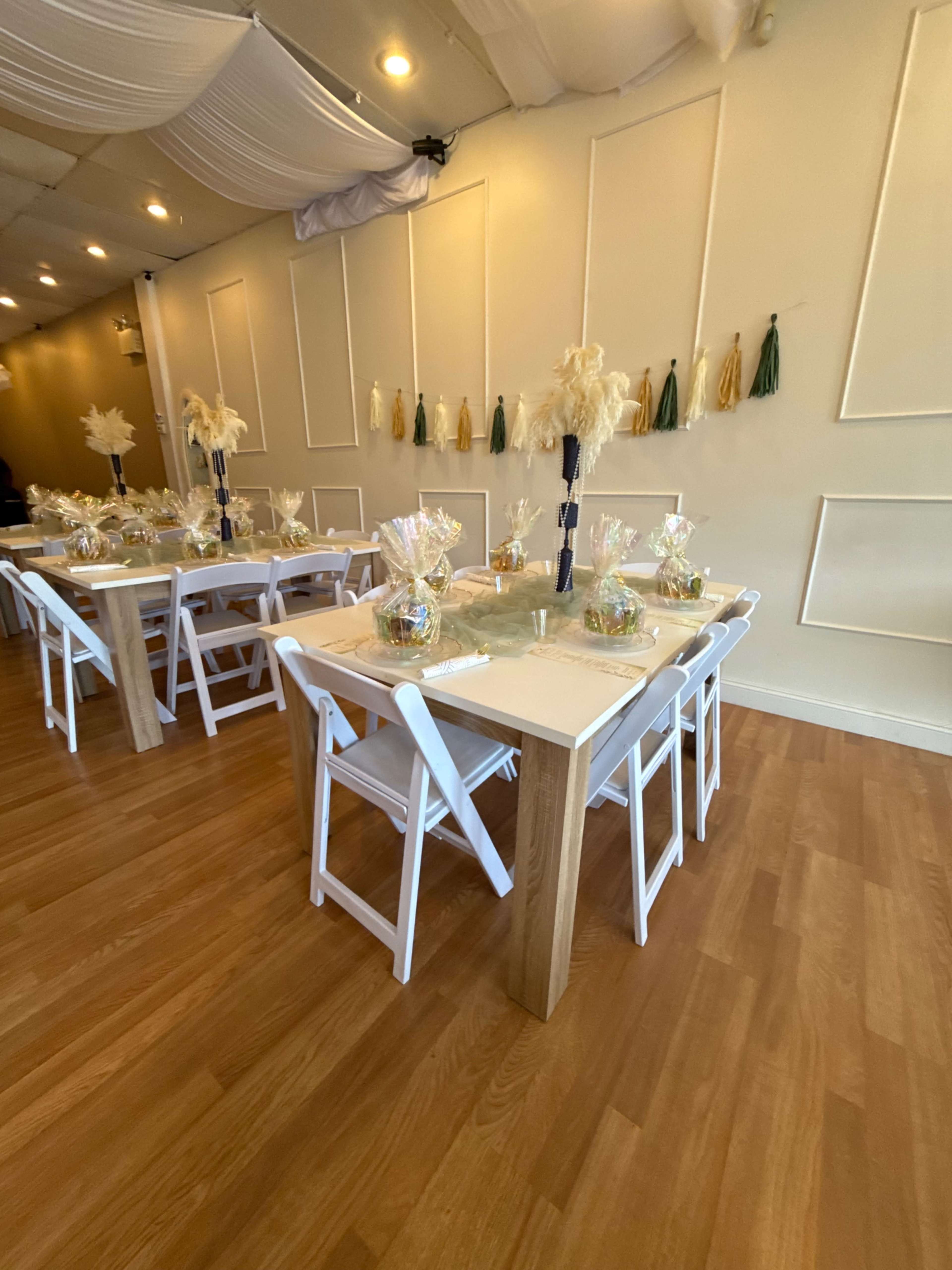 The image shows a decorated event space with several white tables set up, featuring clear bags filled with colorful items, and a wall adorned with decorative tassels.