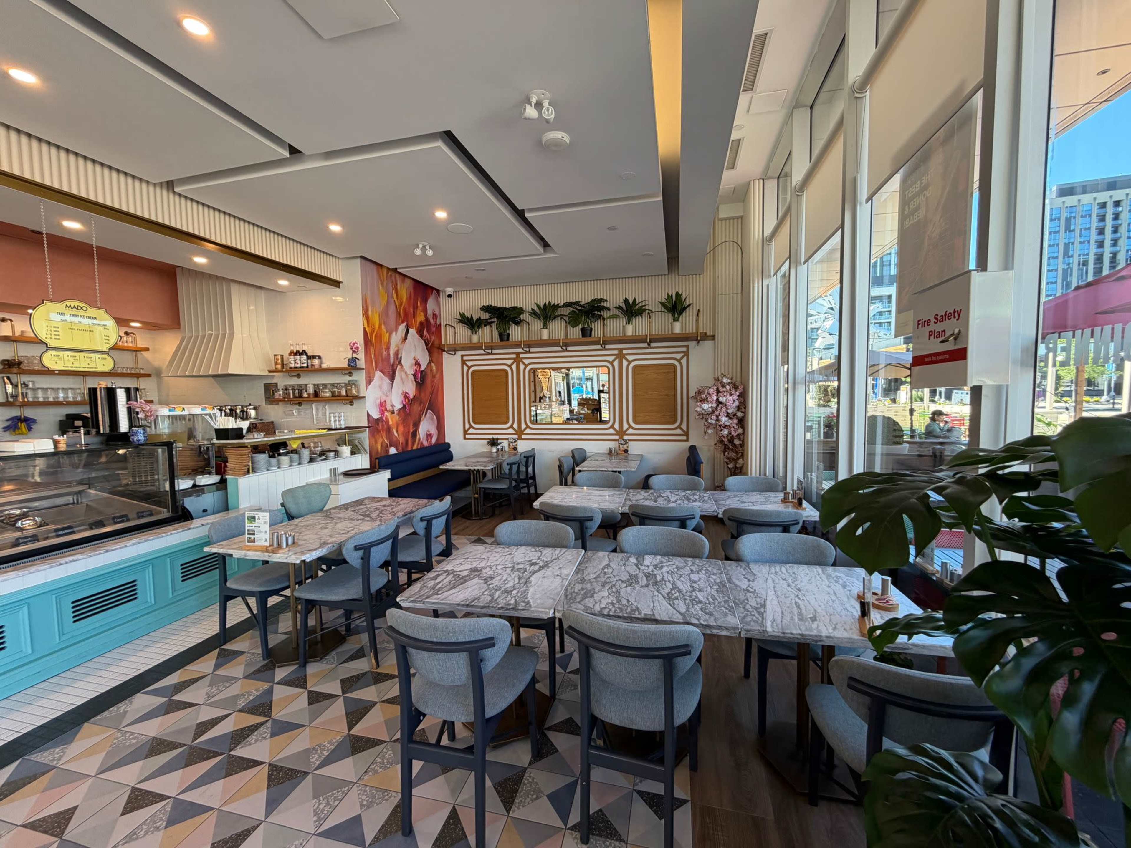 The image shows a modern café interior with marble tables, stylish chairs, and a counter displaying pastries, surrounded by large windows.