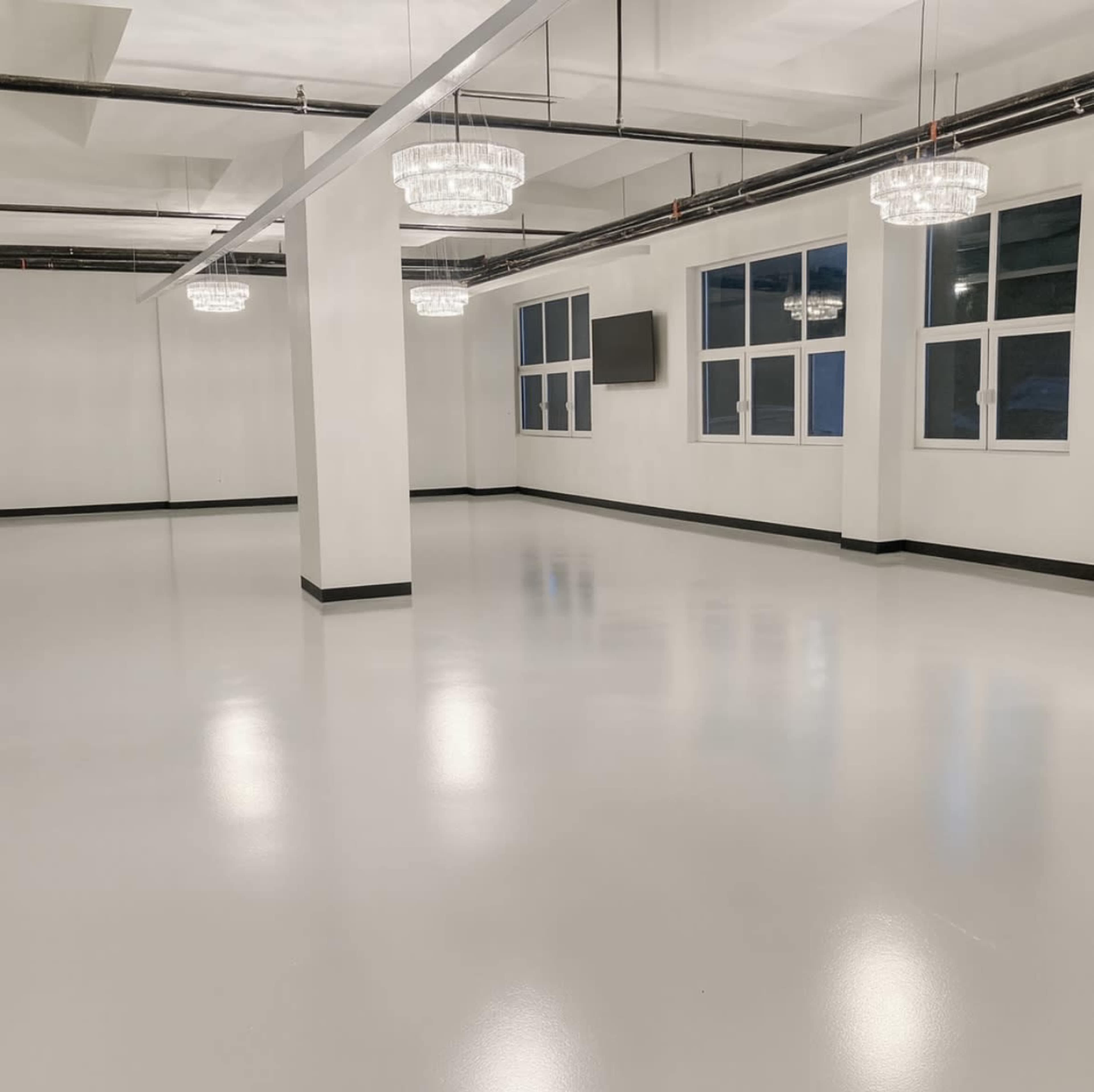 The image shows a spacious, empty room with a polished white floor, several chandeliers, large windows, and a television mounted on the wall.