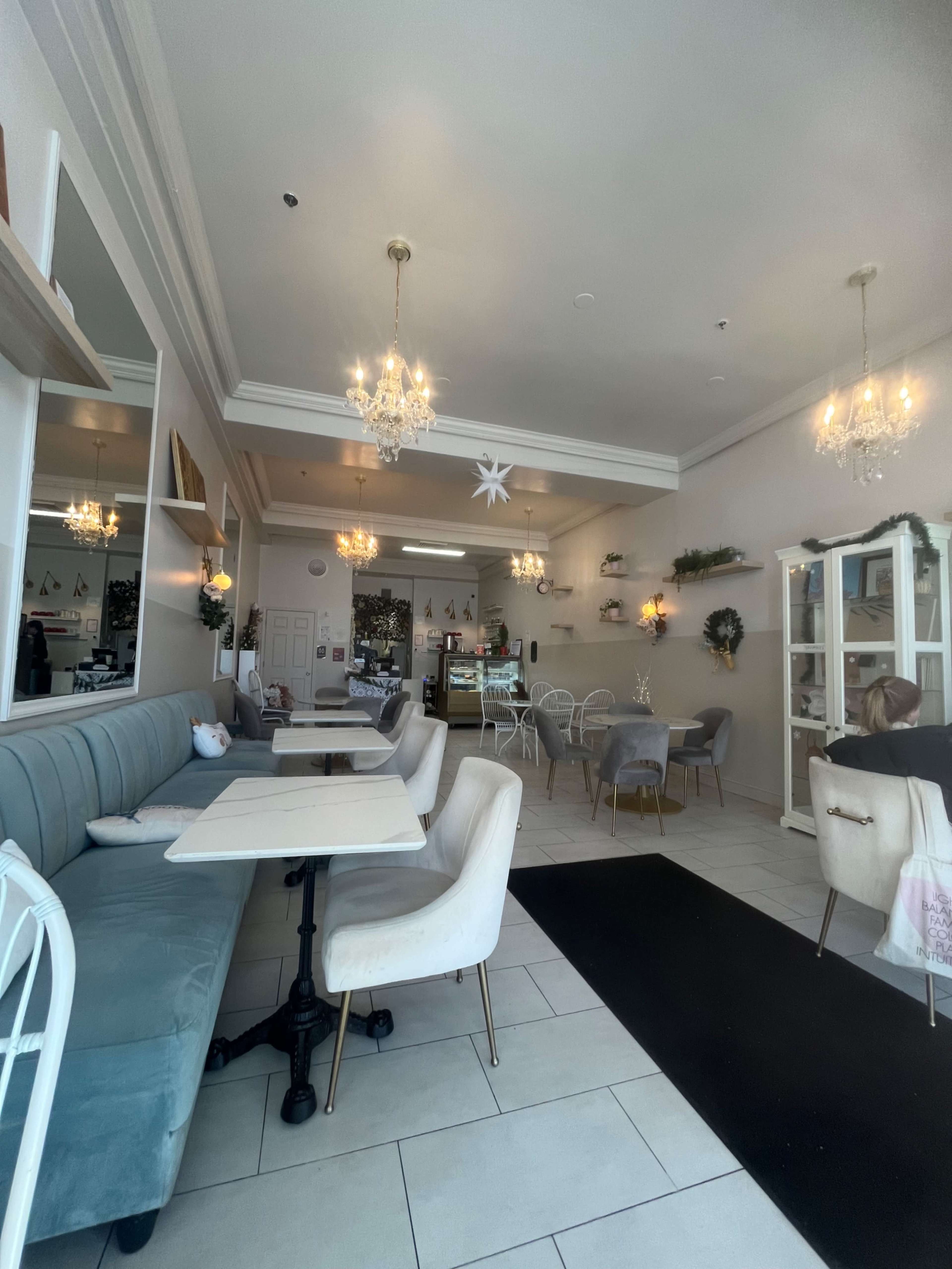 The image shows a clean, well-lit café interior featuring a mix of seating options, including a blue banquette and gray upholstered chairs, with decorative chandeliers and plants.