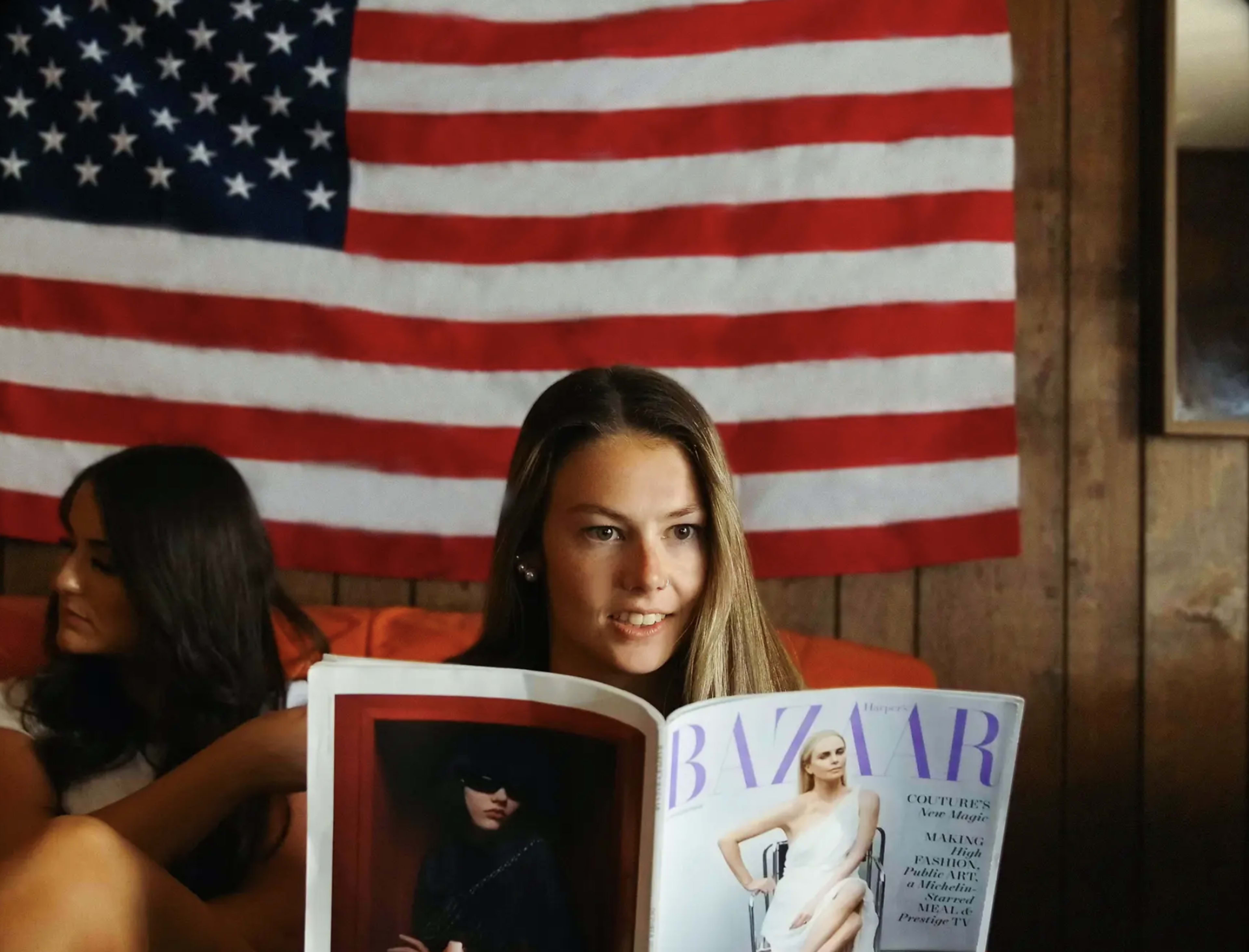 A young woman reads a fashion magazine while sitting against a wall adorned with an American flag.