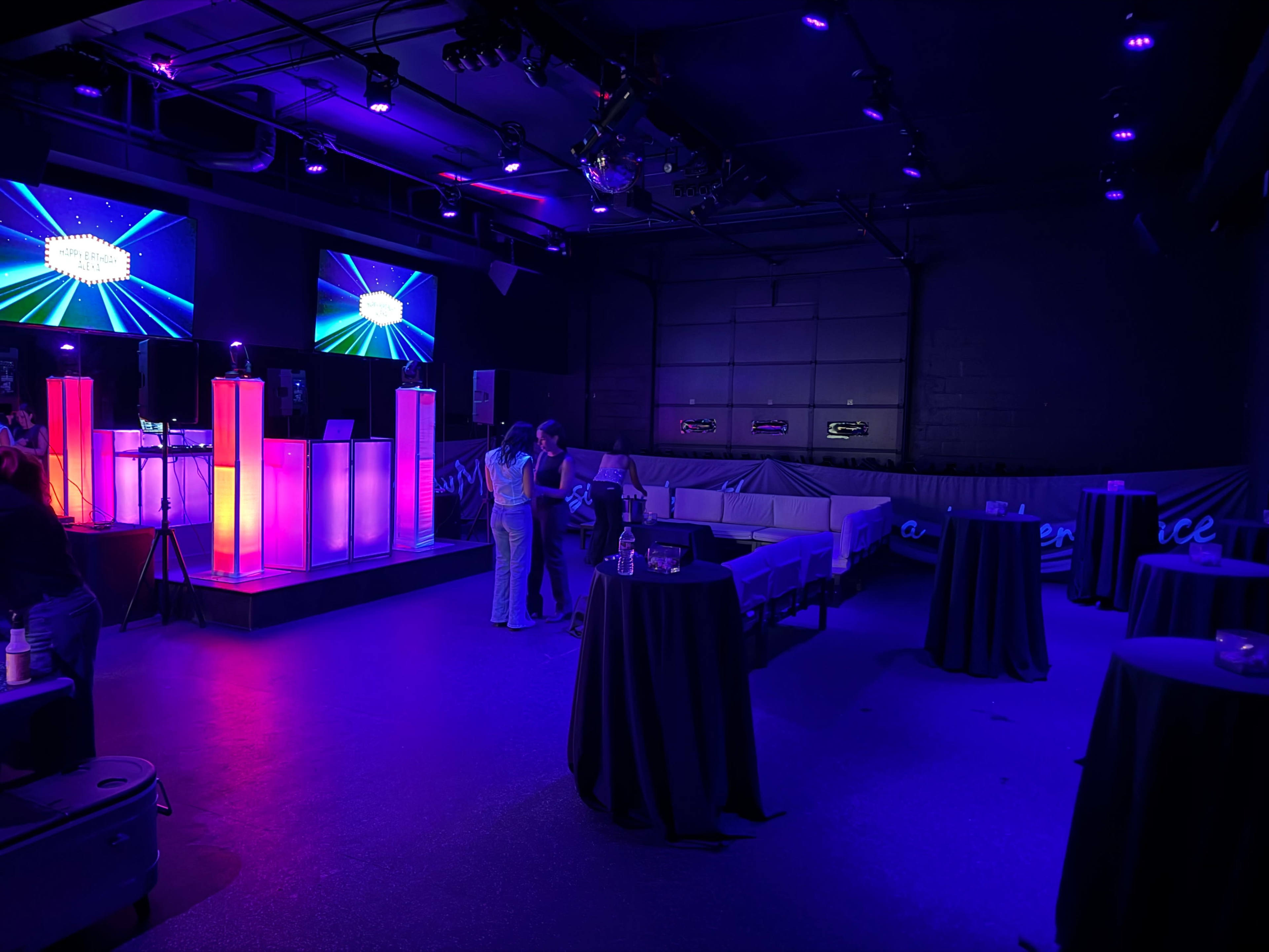 The image shows a modern event space with purple lighting, featuring glowing display stands, two people conversing, and several high tables arranged around the room.