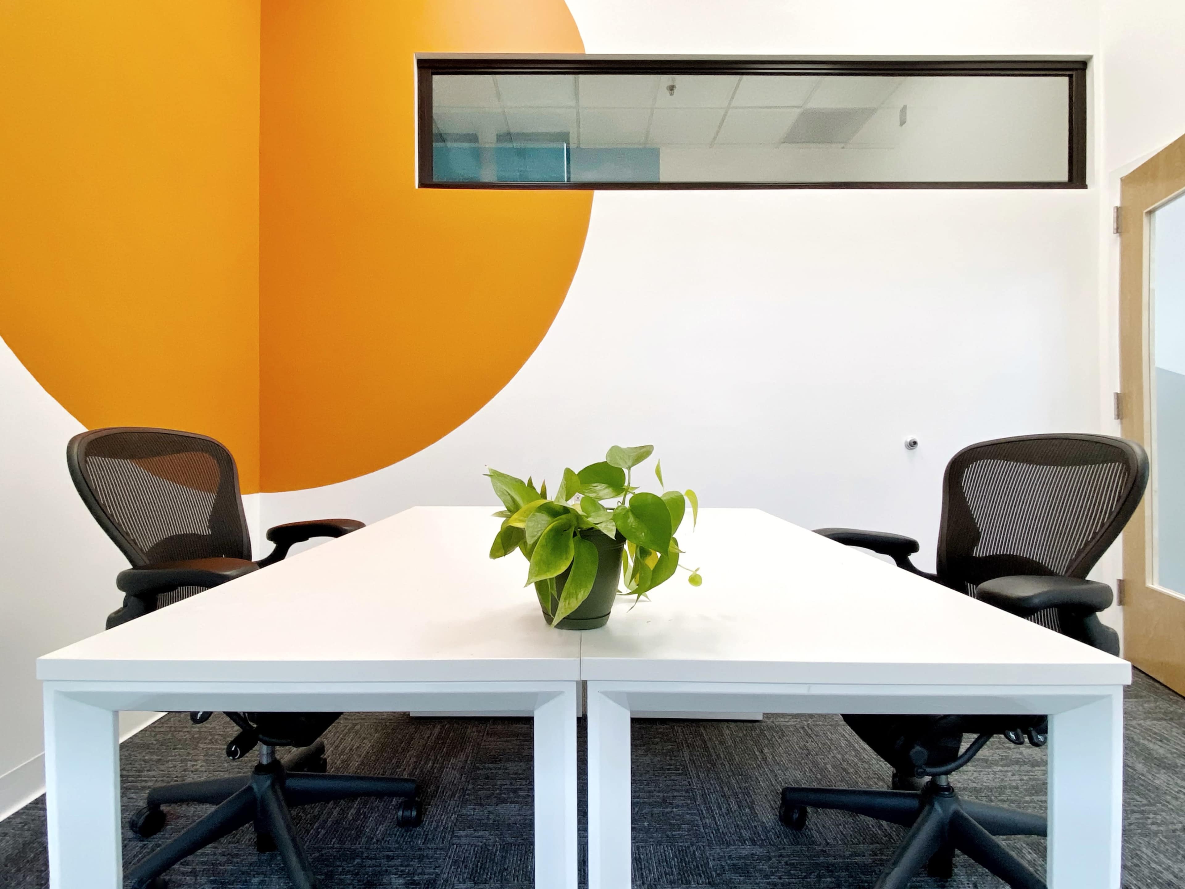 A clean, minimalist office space features a white table with two black chairs and a plant in the center, set against an orange circular accent wall.