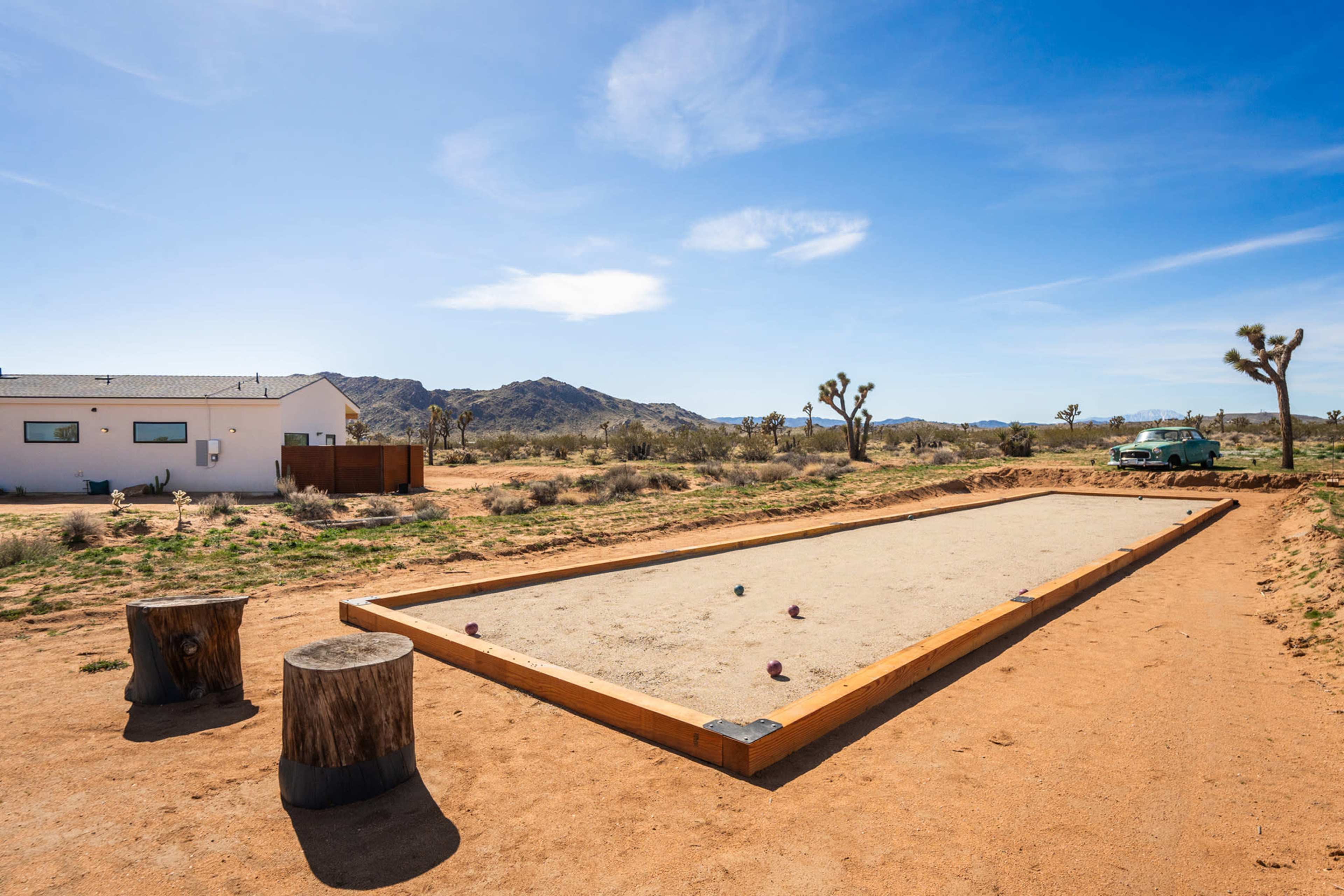 A bocce ball court surrounded by desert landscape and scattered Joshua trees, with an old vehicle visible in the background.