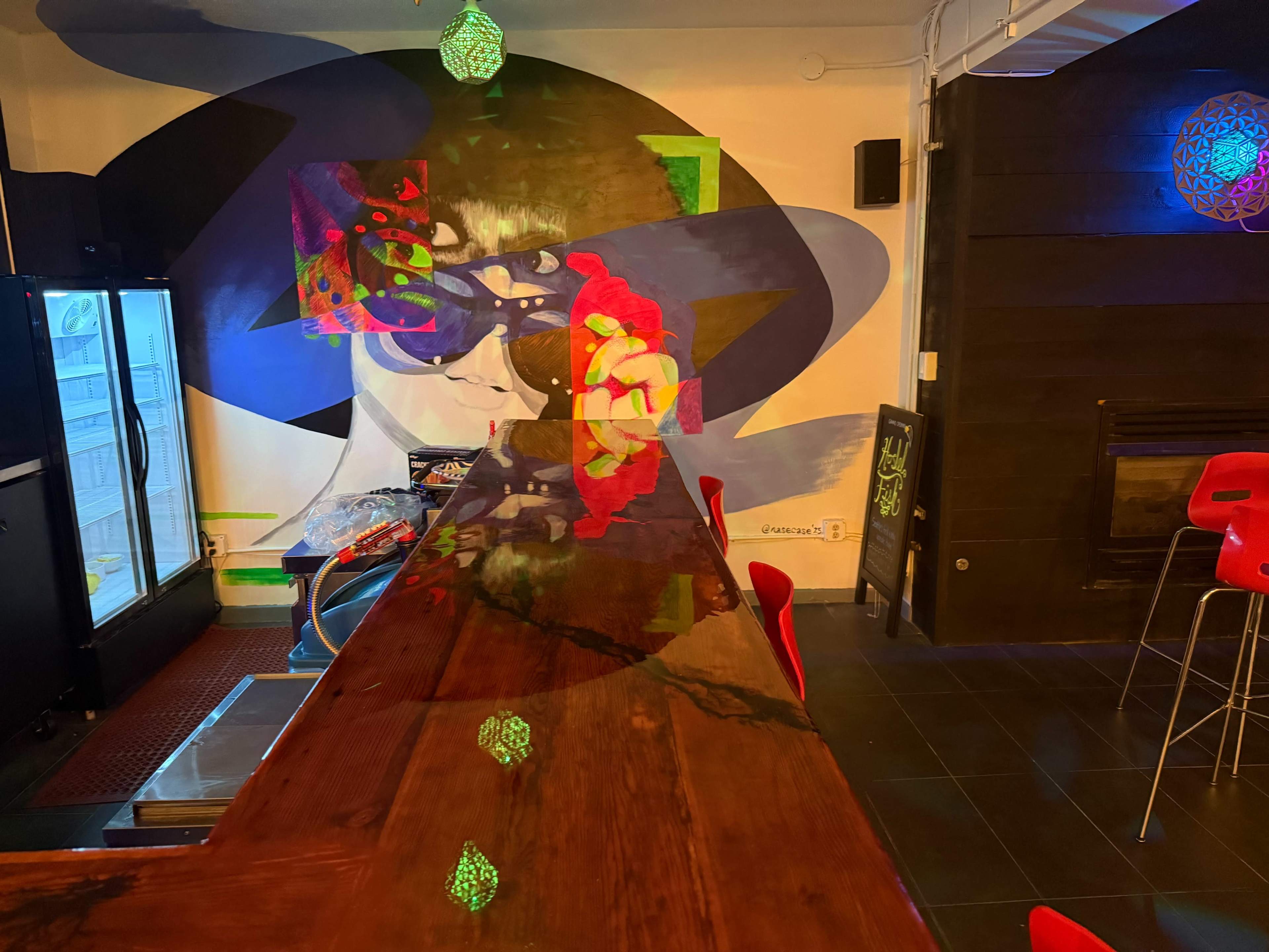 The image shows a vibrant bar area with a wooden counter, colorful wall art featuring a person wearing sunglasses, and illuminated pendant lights above.