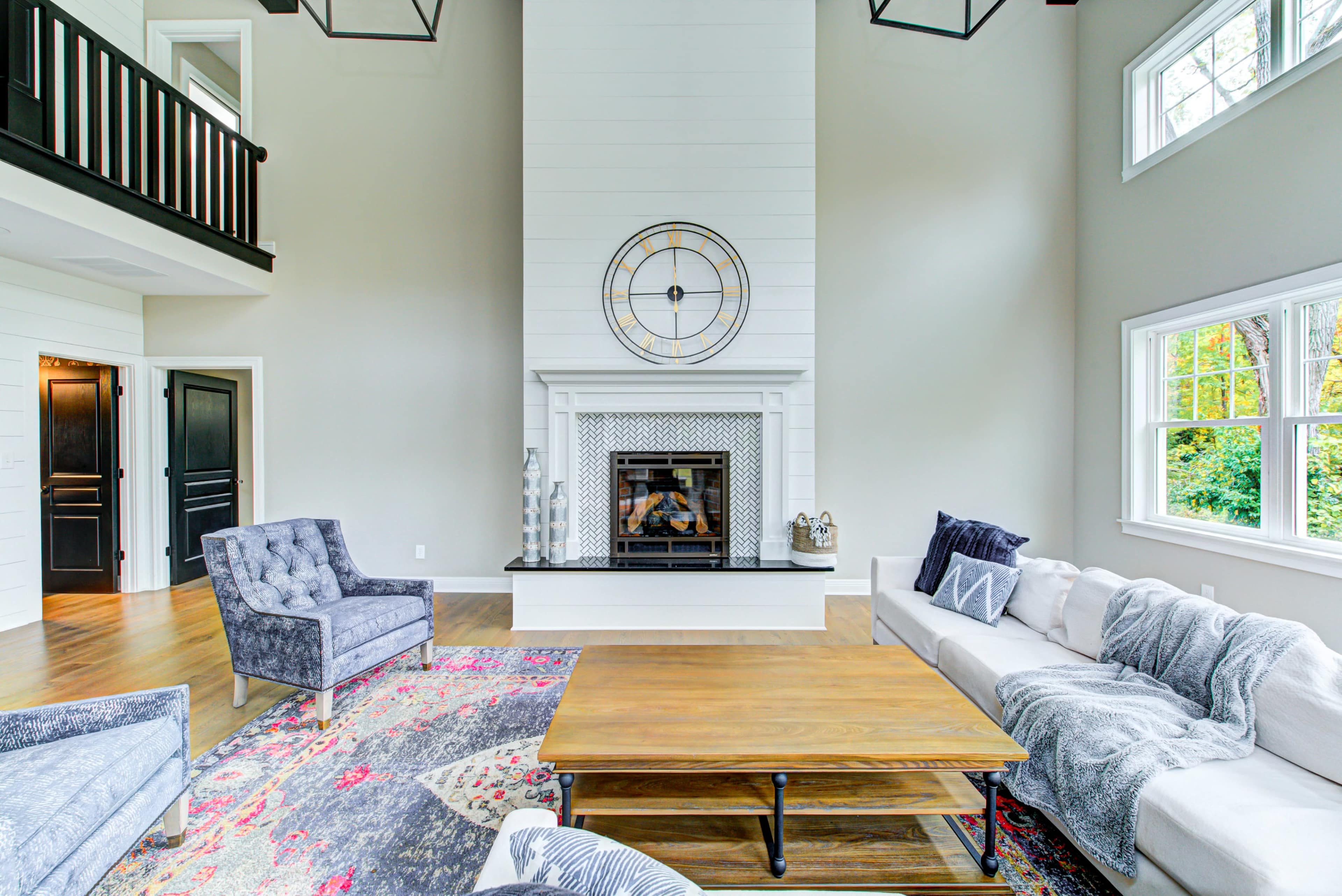 The living room features a large fireplace with a clock above it, surrounded by a sectional sofa and two armchairs, with a wooden coffee table in the center and windows allowing for natural light.