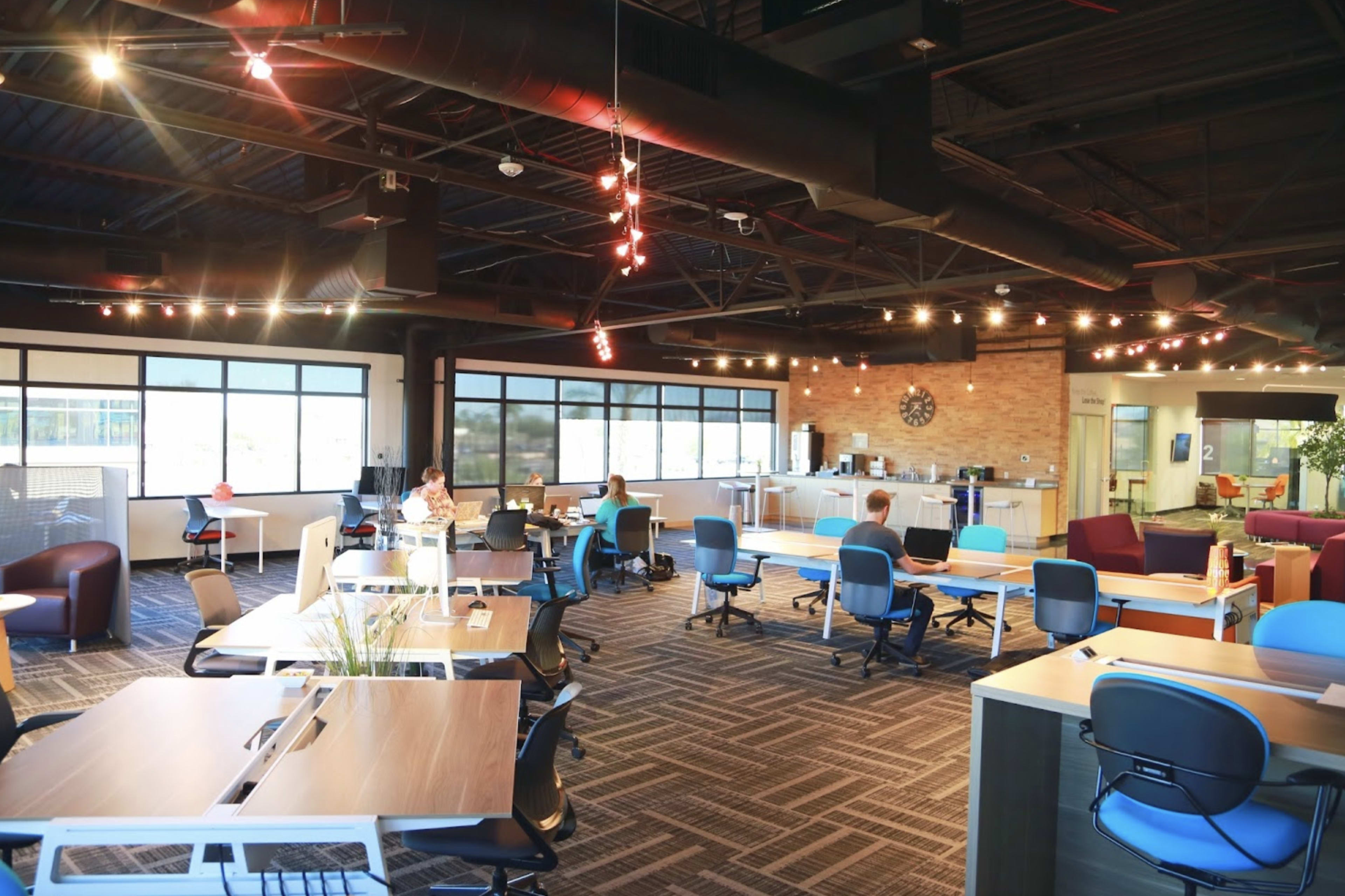 A modern office space with various workstations, a kitchenette, and large windows allowing natural light to enter.