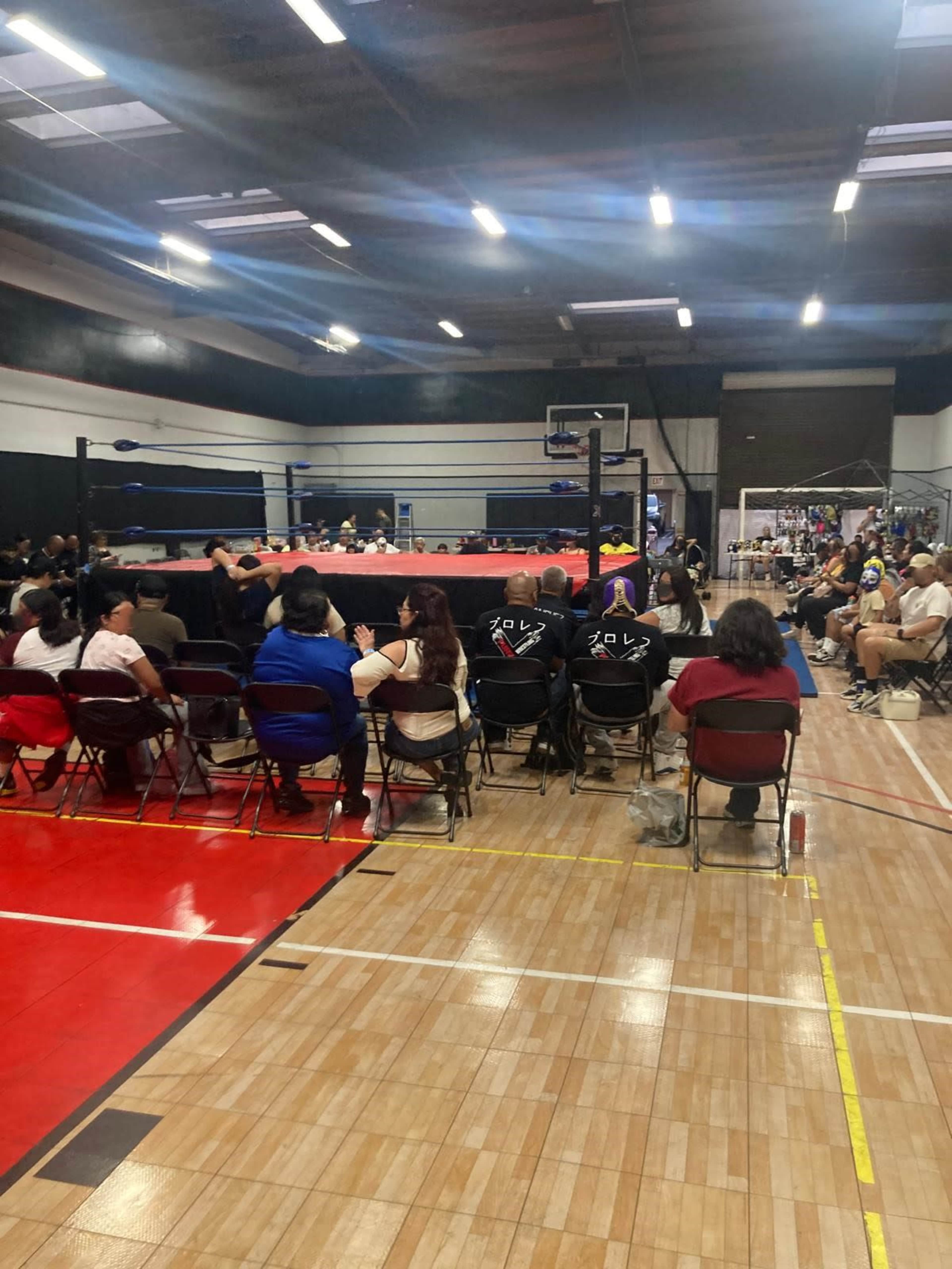A wrestling match is taking place in a gymnasium, with a crowd of spectators seated on folding chairs surrounding the ring.