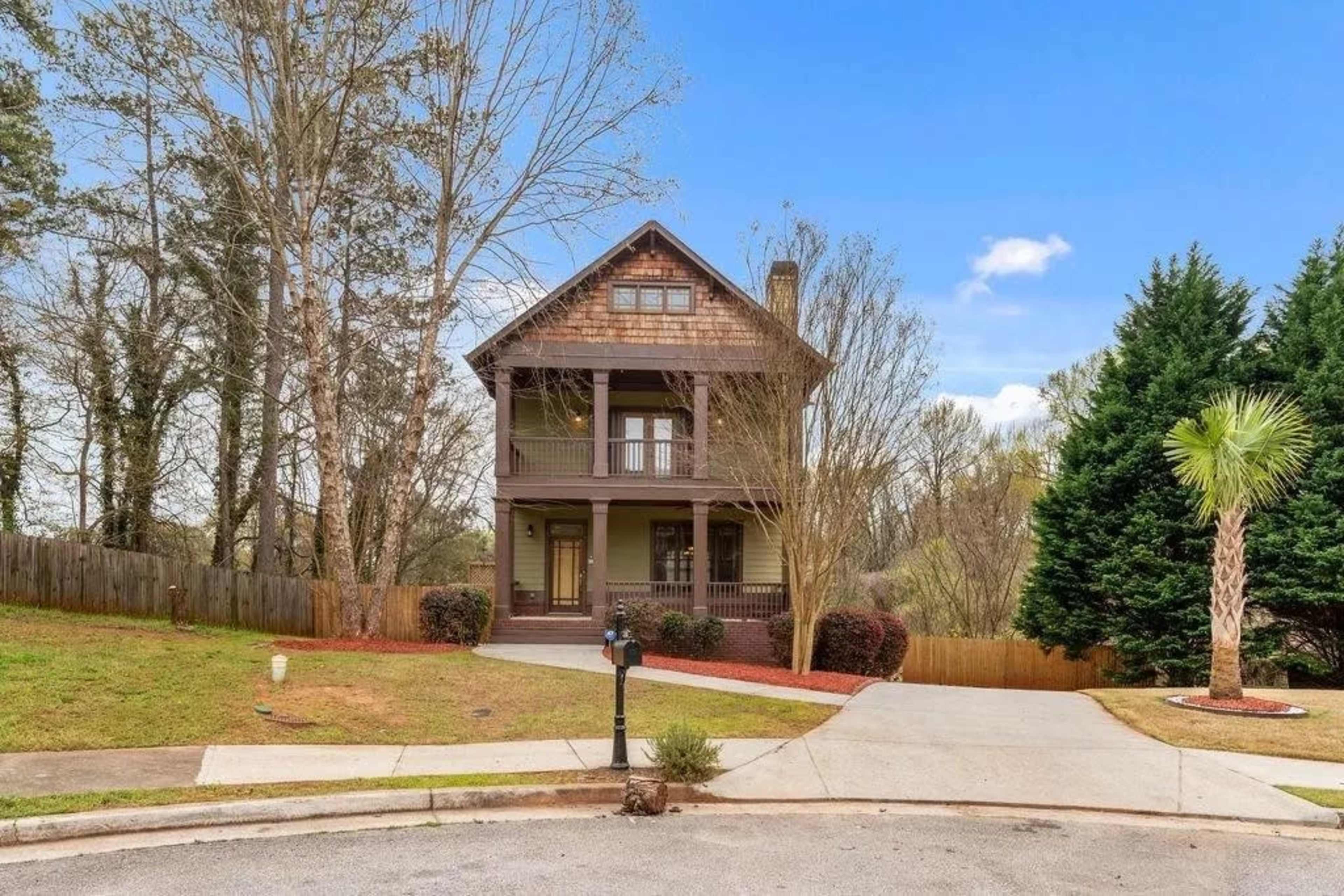 The image shows a two-story house with a wooden exterior, situated at the end of a driveway, surrounded by trees and shrubs.