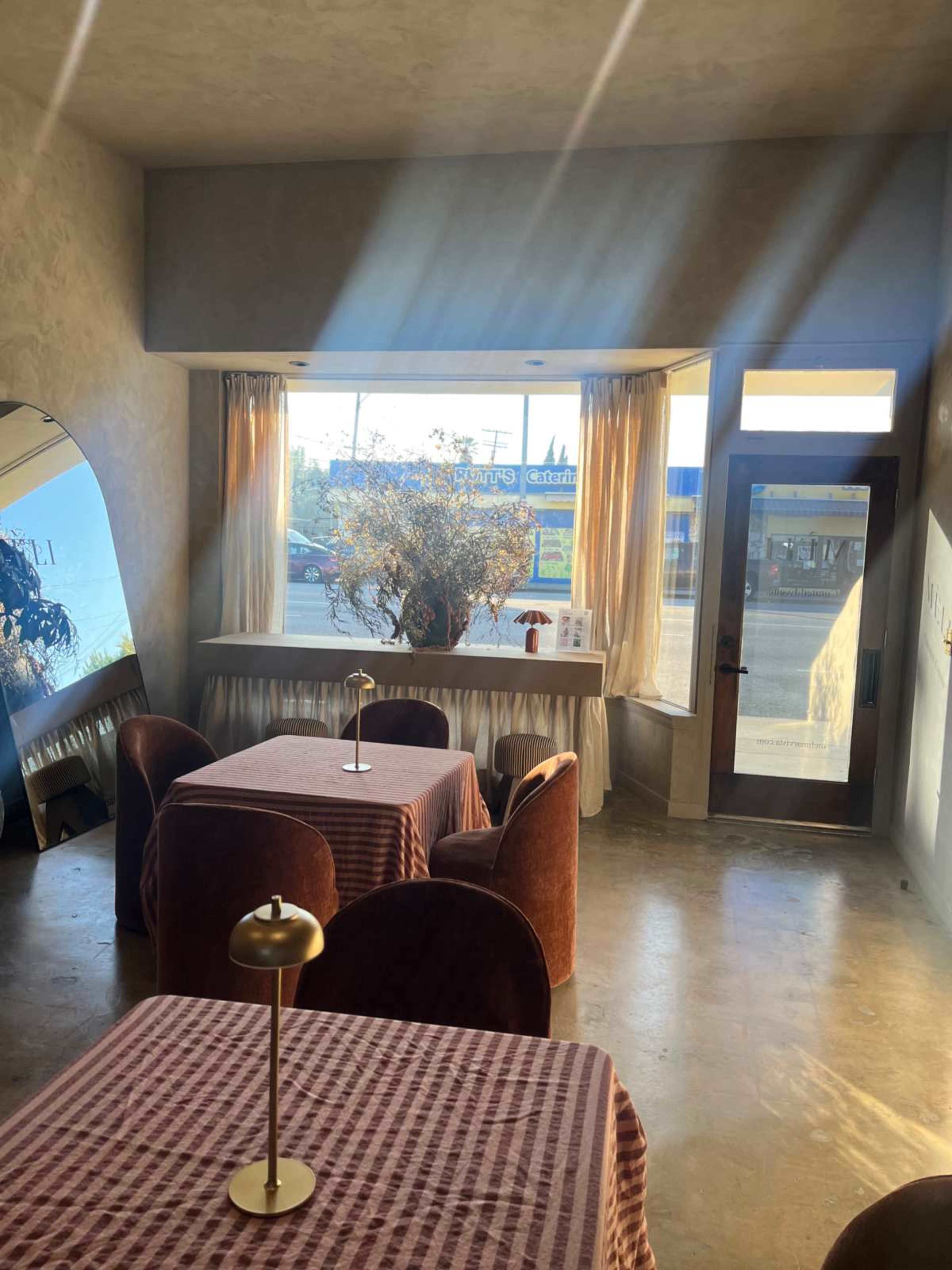 The image shows a minimalist restaurant interior featuring two tables with red checkered tablecloths, a large mirror, and a window with natural light illuminating the space.