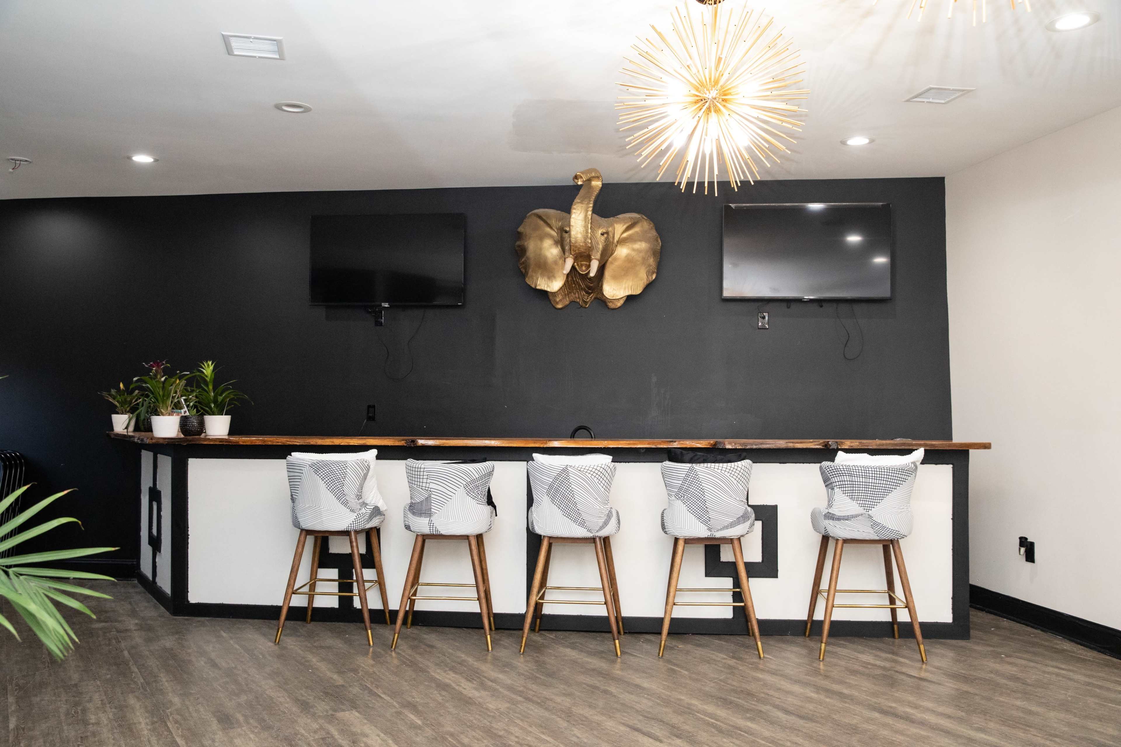 A modern bar area with a wooden counter, five stools, two wall-mounted televisions, and a decorative wall piece resembling an elephant's head.